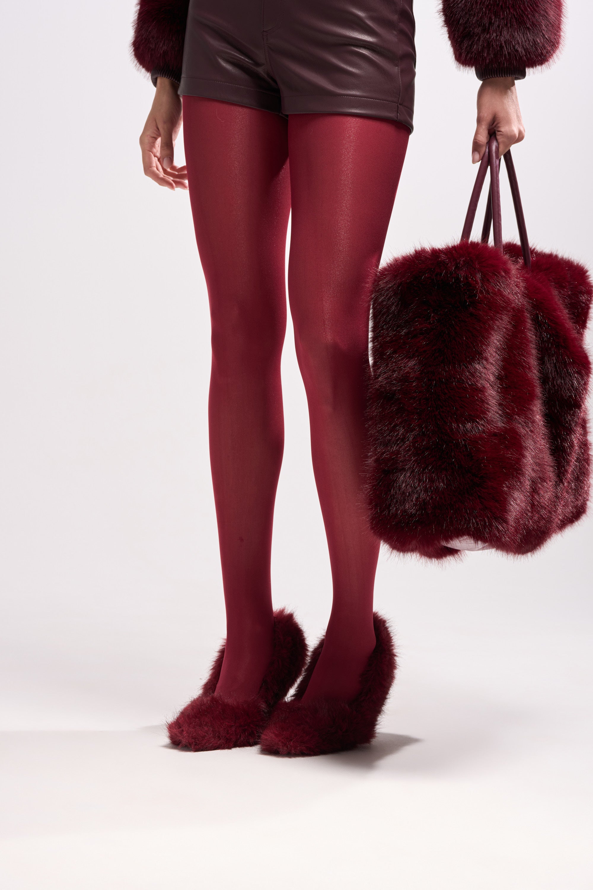 A person stands against a white background wearing OXBLOOD OPAQUE TIGHTS, leather shorts, a furry jacket, furry shoes, and holding a matching red furry handbag—ideal for an autumn wardrobe.