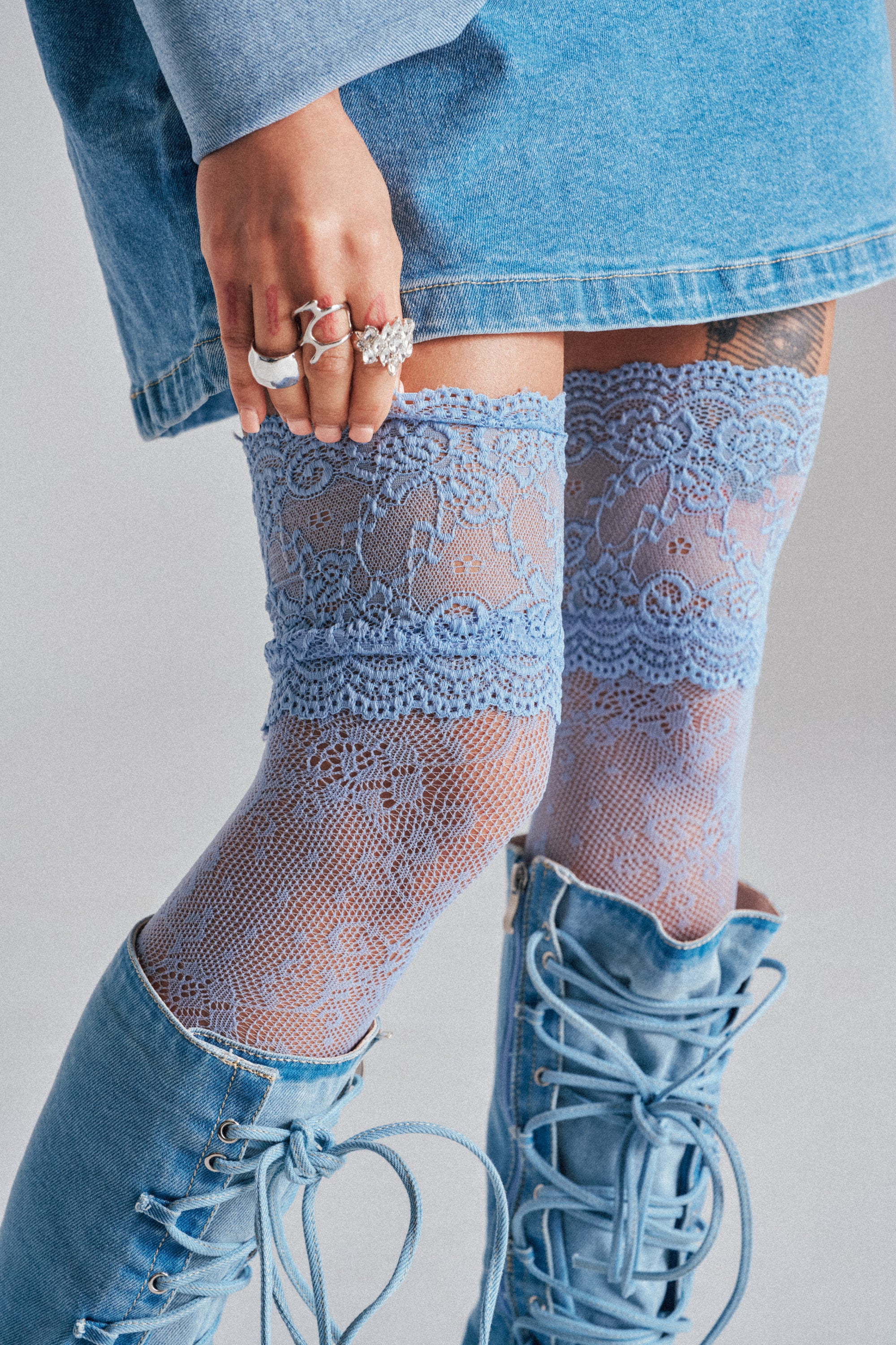 A person in a denim skirt, CLASSY VIBES THIGH HIGH STOCKINGS in pastel blue, and knee-high denim boots. Their hand with multiple rings rests on their thigh. The color palette features mainly shades of blue.