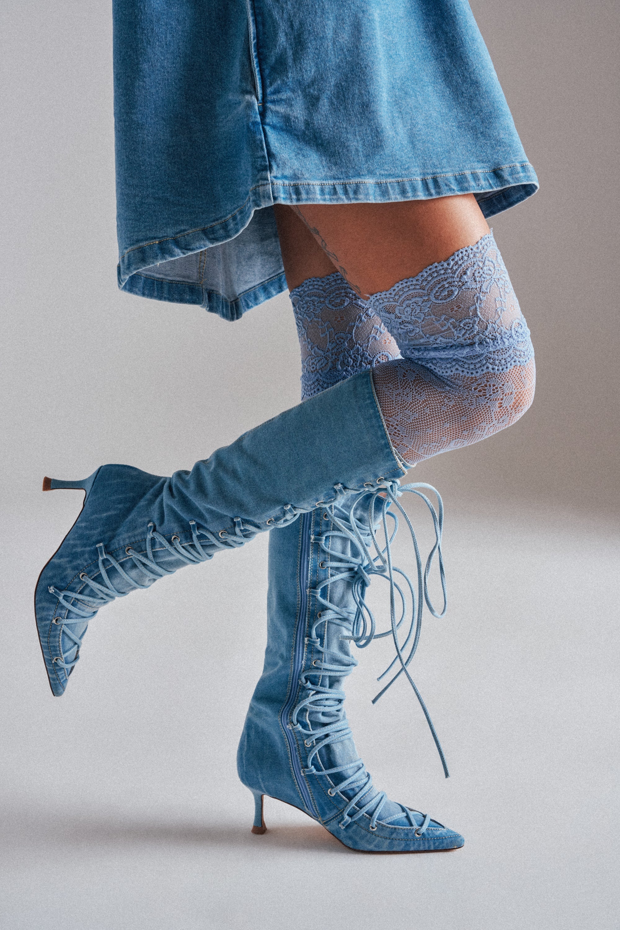 A person wearing a denim dress, CLASSY VIBES THIGH HIGH STOCKINGS in pastel blue, and blue lace-up denim heeled boots stands in profile with one leg slightly bent against a plain background. 1