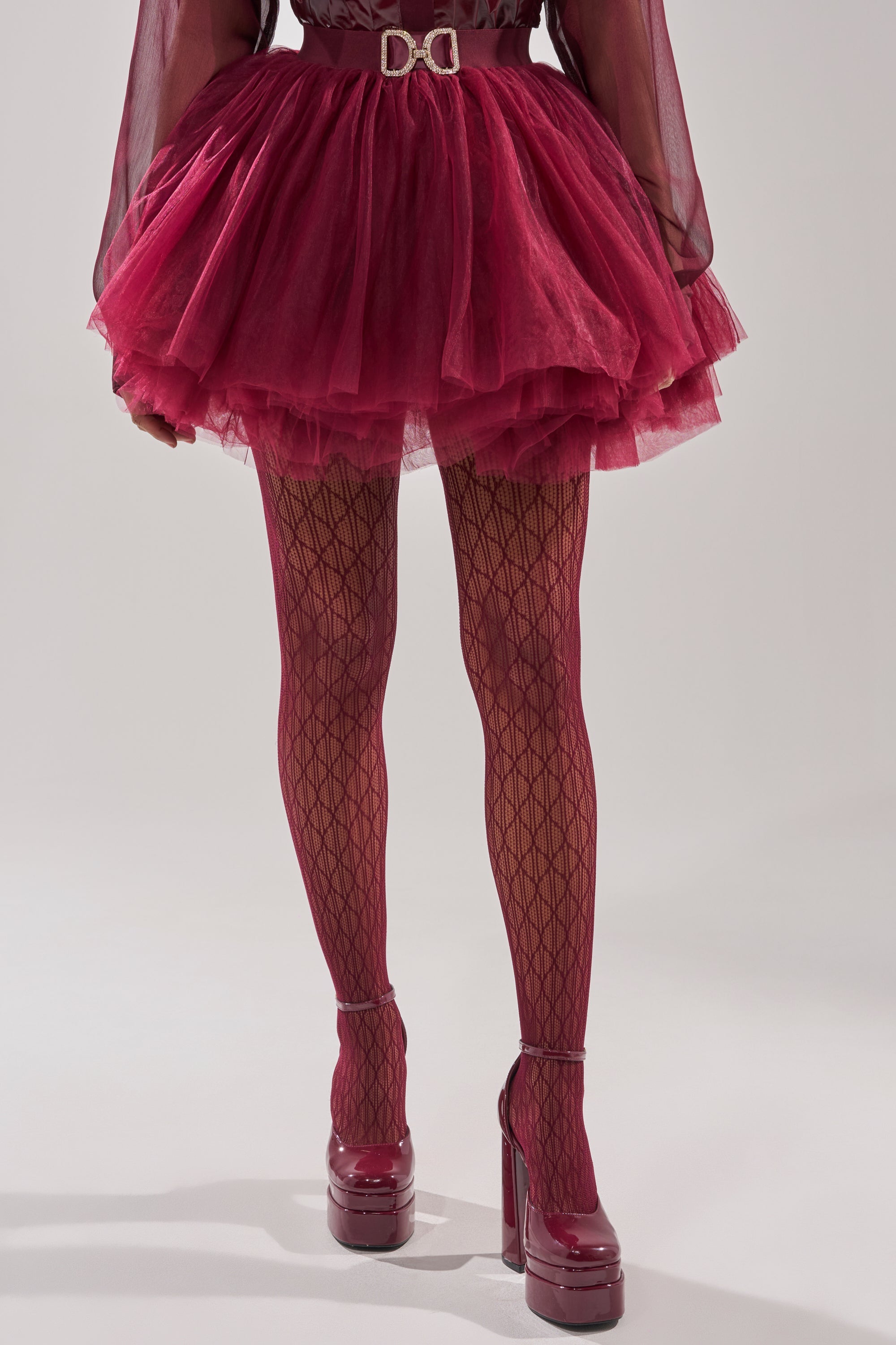 Wearing BELLA TIGHTS with a lace diamond pattern, a burgundy tulle mini skirt, sheer top, platform heels, and a belt with a bold buckle. The background is plain and light-colored. 3