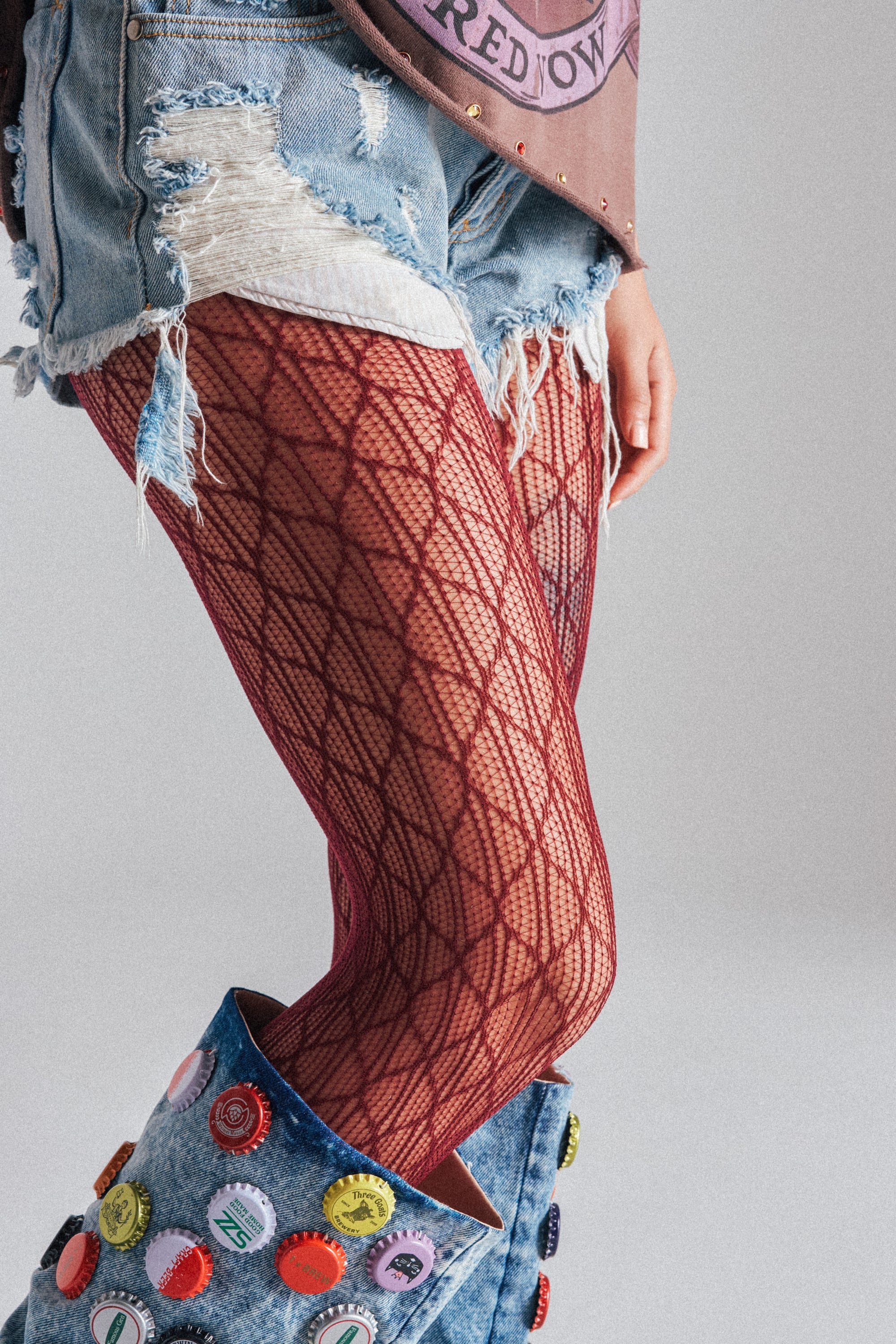 A person’s lower half is shown against a plain background, wearing ripped denim shorts, denim boots with colorful buttons, and red BELLA TIGHTS featuring a patterned fishnet design. 1