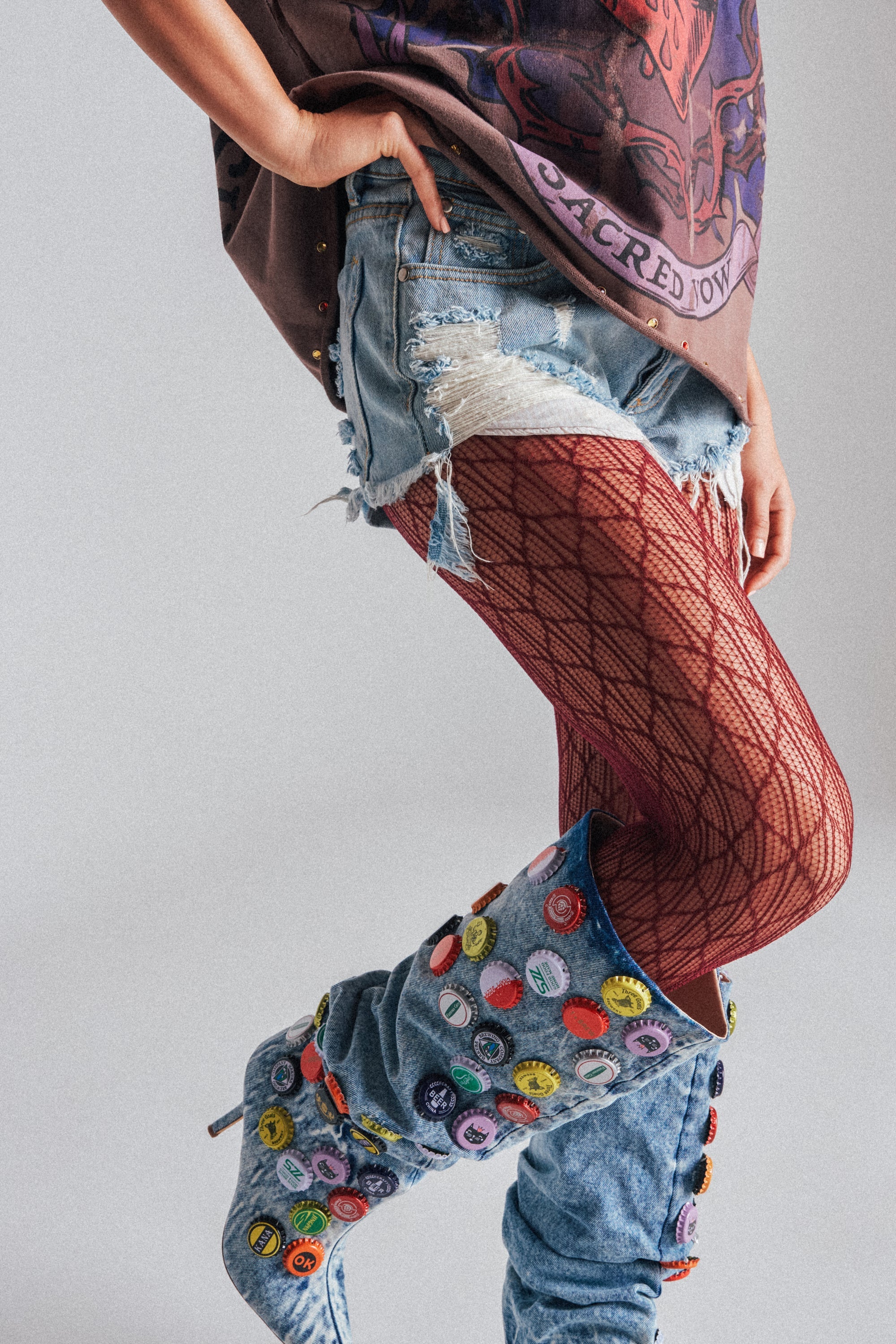 Only the lower body is visible—a person stands with one hand on their hip, wearing ripped denim shorts, denim boots adorned with colorful pins and buttons, and BELLA TIGHTS in a bold red pattern. 2