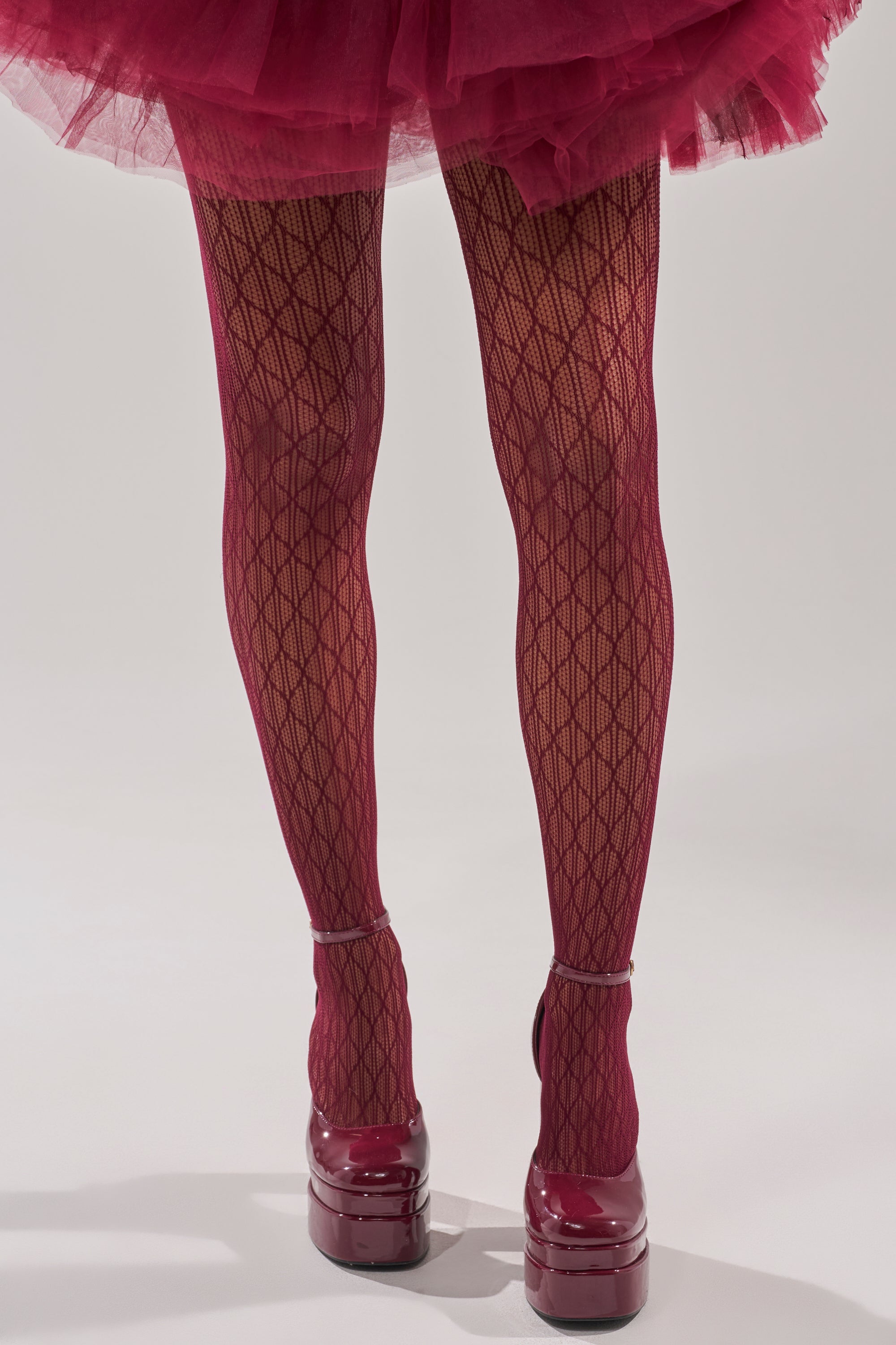 A person wearing BELLA TIGHTS with a lace diamond pattern, a matching burgundy tulle skirt, and glossy platform heels stands with their back to the camera against a plain light background. 4