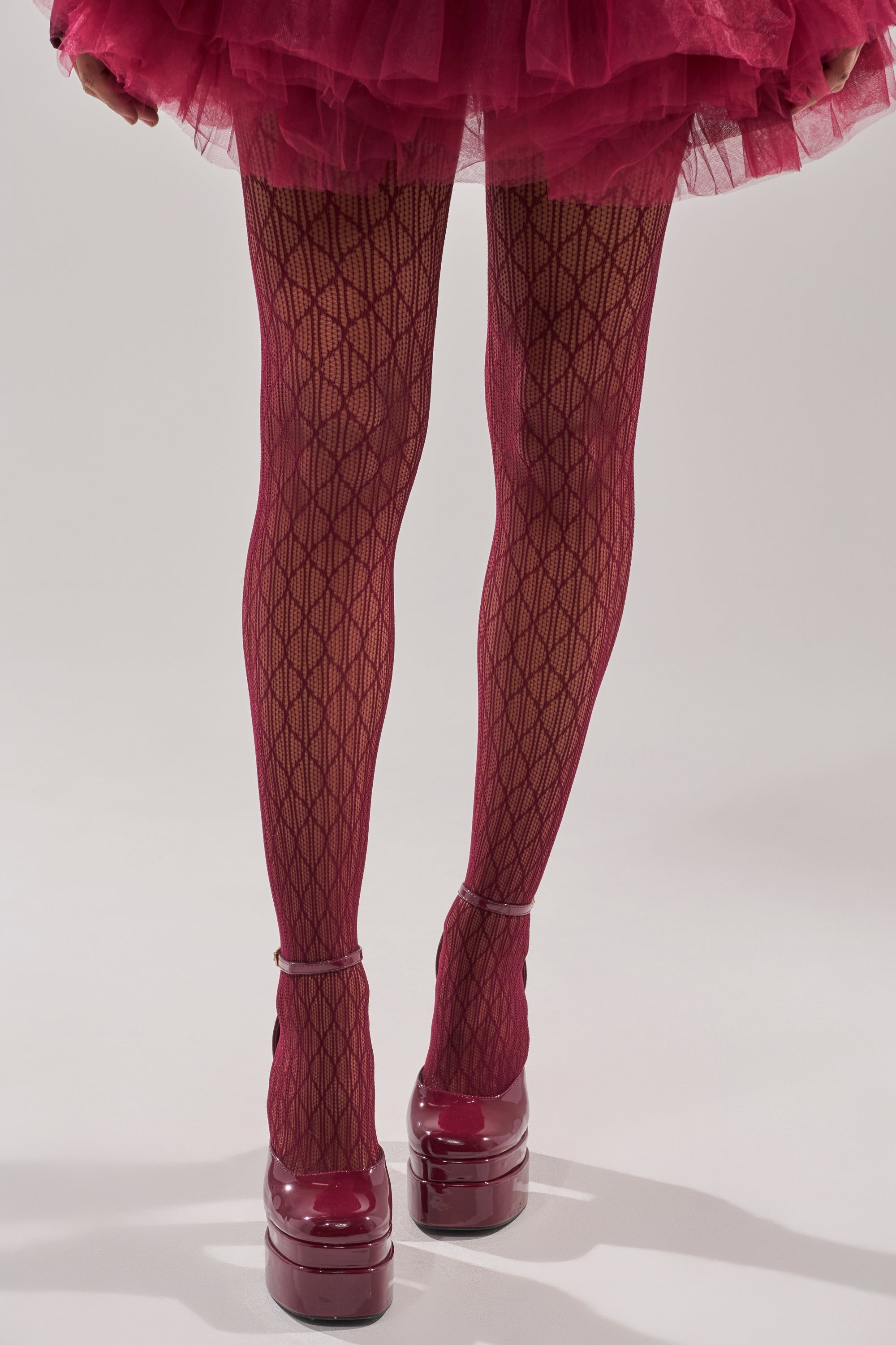 Wearing BELLA TIGHTS in patterned black fishnet, a person pairs them with a magenta tulle skirt and shiny platform heels, standing before a plain background. Cropped at the knees, the look offers perfect holiday party tights inspiration. 5