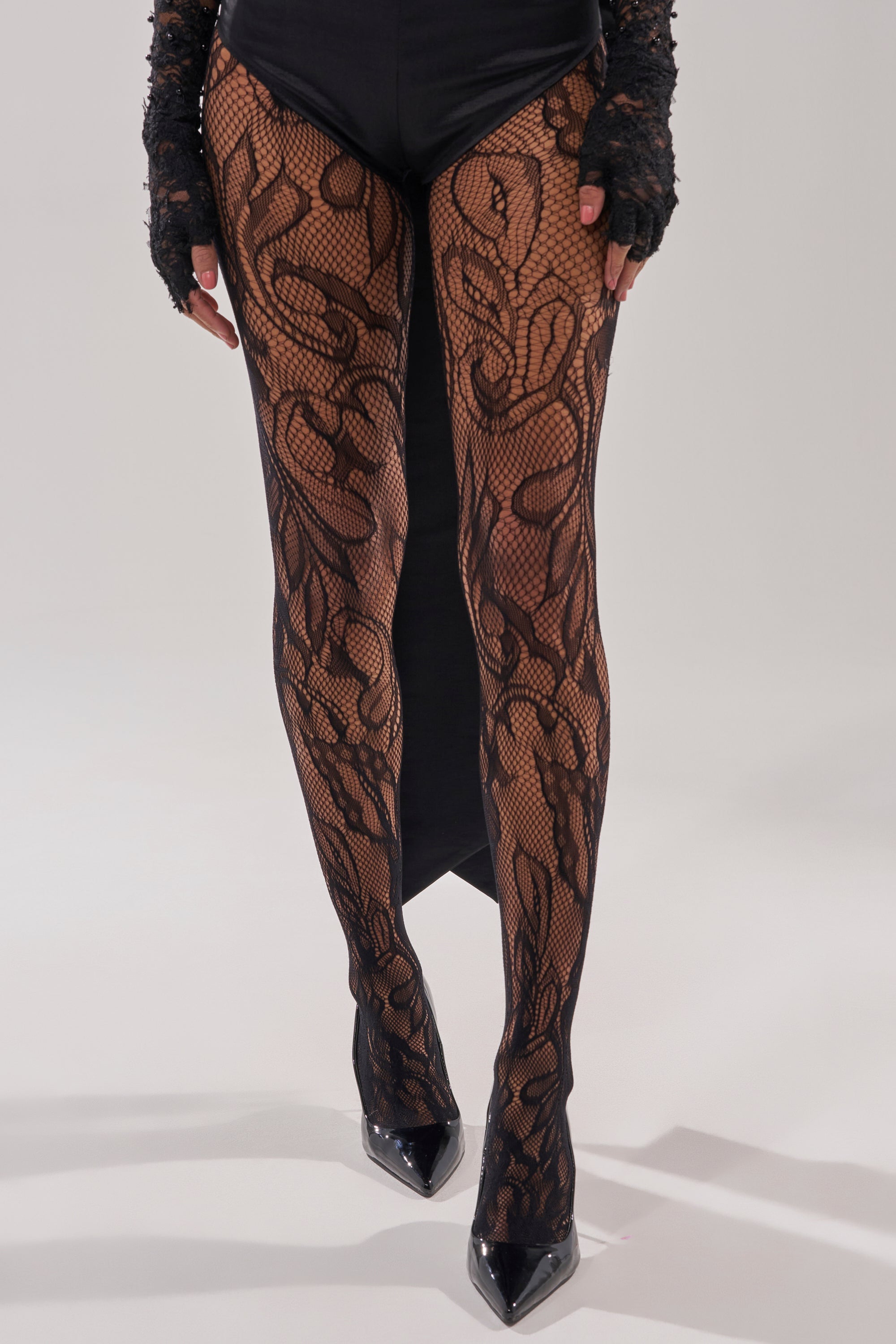 A person stands against a plain light background wearing LILI TIGHTS with a lacelike floral pattern, paired with black gloves and black high-heeled shoes. 1