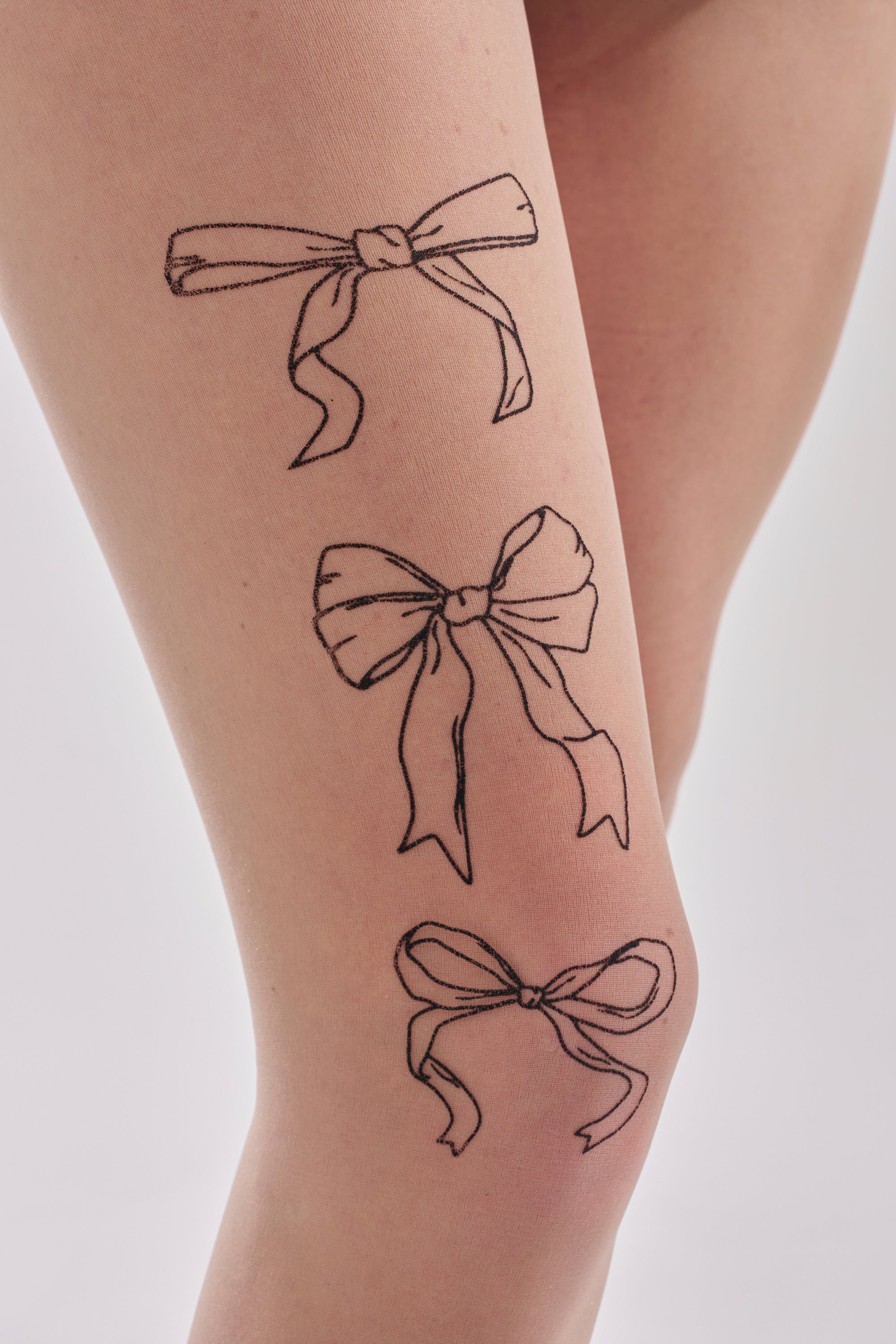 Three black line art bows of varying sizes and styles are drawn on a light-skinned thigh, resembling the delicate patterns of WRAP WITH A BOW TIGHTS. The plain, light background highlights the elegant design. 4