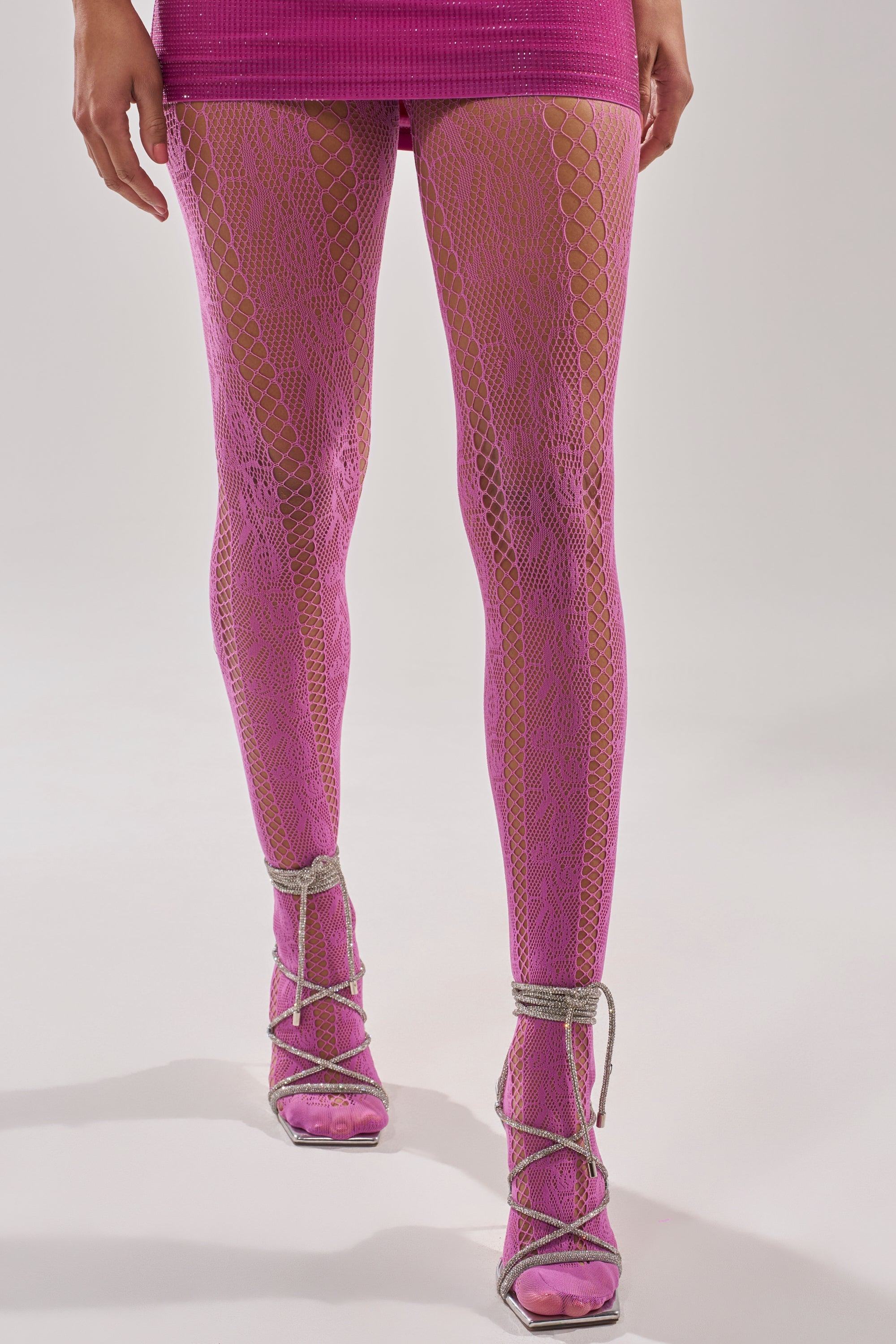 A person stands against a plain background, showcasing FEMME TIGHTS IN PINK with a geometric pattern, paired with a matching skirt and silver strappy high-heeled sandals for a bold, trendy look. 0