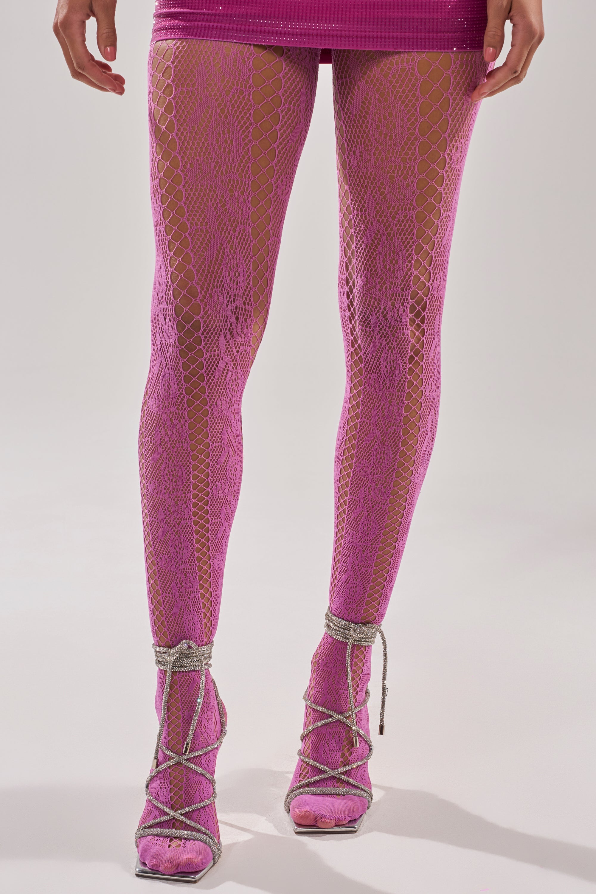 A person models FEMME TIGHTS IN PINK with a matching skirt and silver strappy high heels, standing against a plain light background. 1