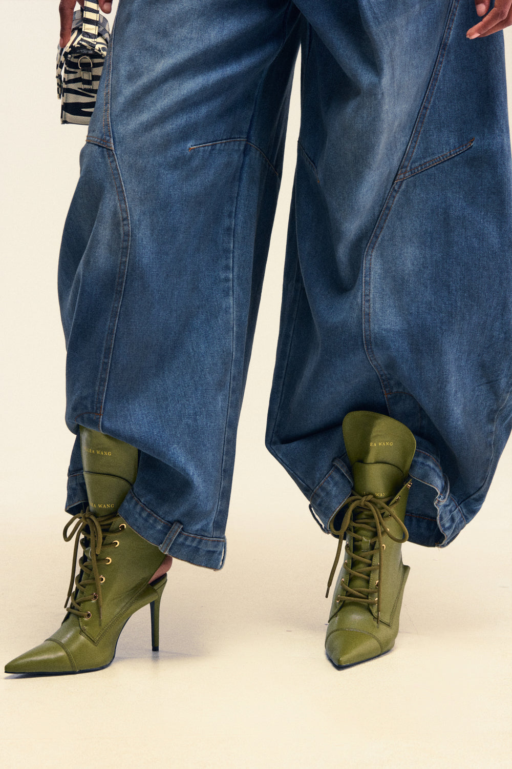 A person wears wide-legged denim pants paired with the AZALEA WANG XIOMARA Olive Lace Up Stiletto Booties and holds a small black and white patterned handbag, with only their legs and lower body visible.