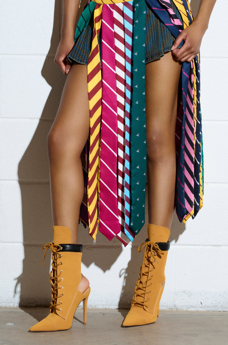 A person is wearing AZALEA WANG XIOMARA Yellow Lace Up Stiletto Booties and a skirt made from colorful neckties. The mid-thigh shot highlights their unique style against a stark white brick wall.