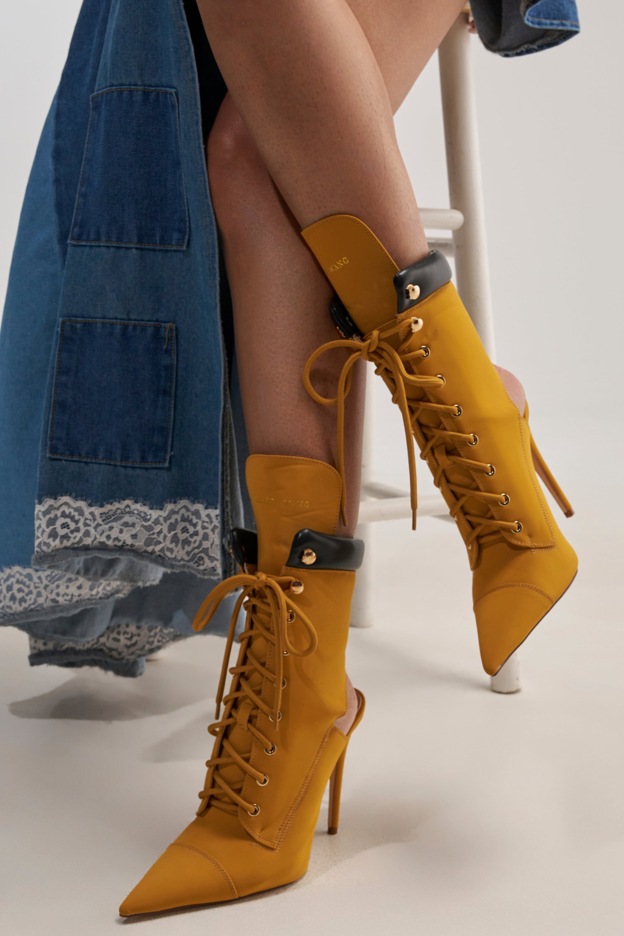 Wearing the AZALEA WANG XIOMARA YELLOW LACE UP STILETTO BOOTIE, a person sits on a white stool in a long blue denim skirt with lace trim. The vegan leather booties feature pointed toes, stiletto heels, black collars, and chic lace-up detailing.