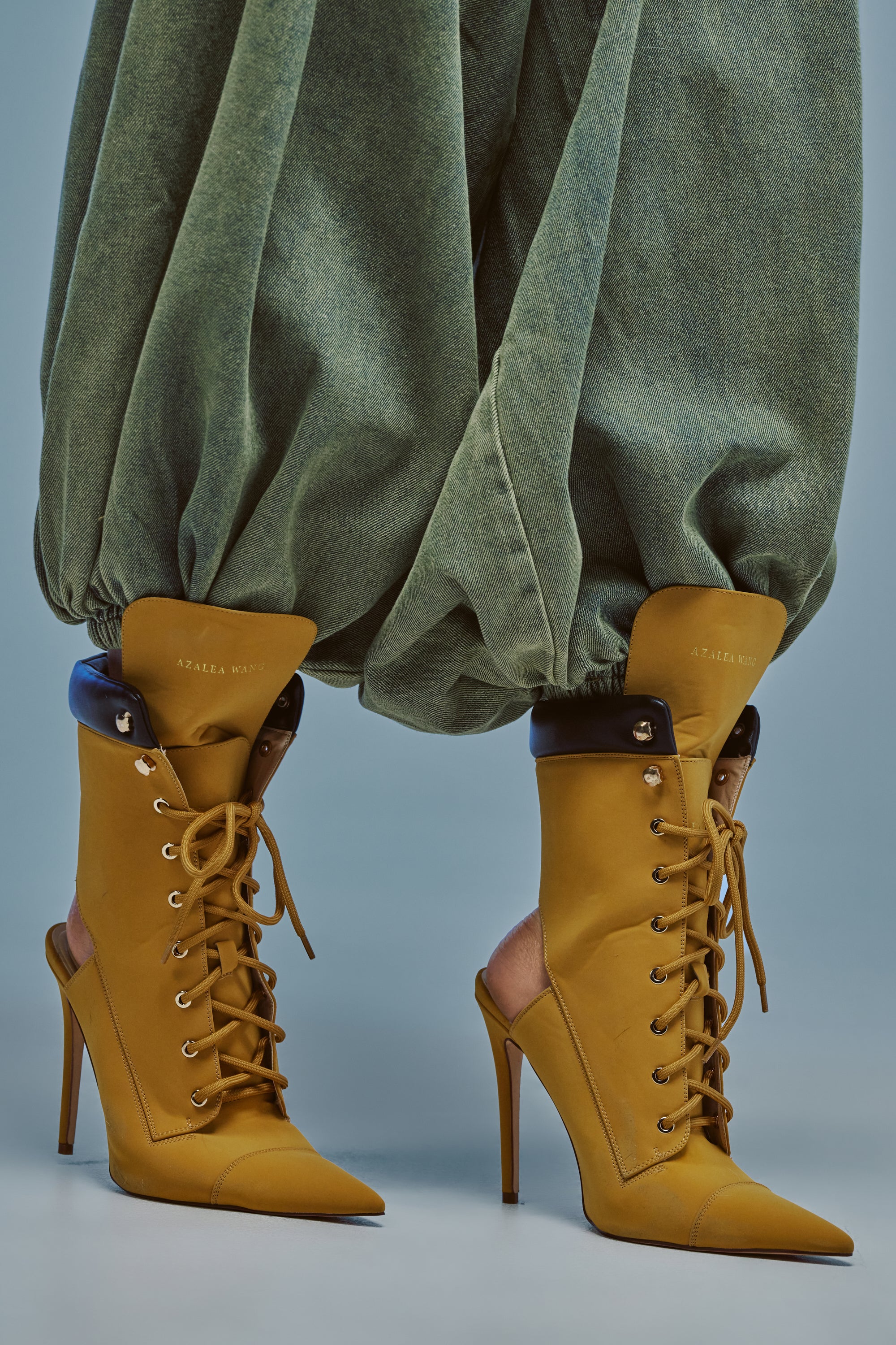 A person wearing loose, green, cinched pants paired with the AZALEA WANG XIOMARA YELLOW LACE UP STILETO BOOTIE, which features pointed toes and black trim at the top. The background is a solid, light grayish blue.