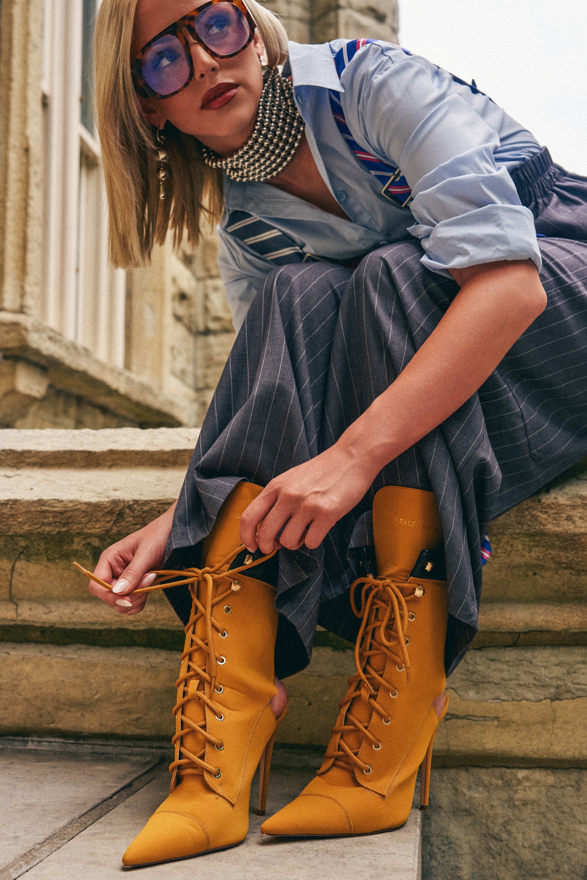 A fashionable woman with blonde hair, large sunglasses, and pearl jewelry ties the laces of her AZALEA WANG XIOMARA YELLOW LACE UP STILETTO BOOTIE while sitting on stone steps, styled in a blue shirt and gray pinstripe skirt for a statement look.