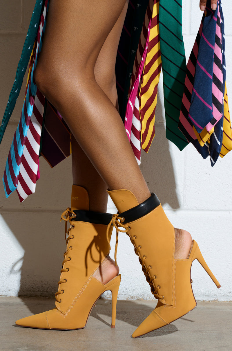 Someone wearing AZALEA WANG XIOMARA yellow lace-up stiletto booties with black trim against a backdrop of colorful striped ties hanging from above.