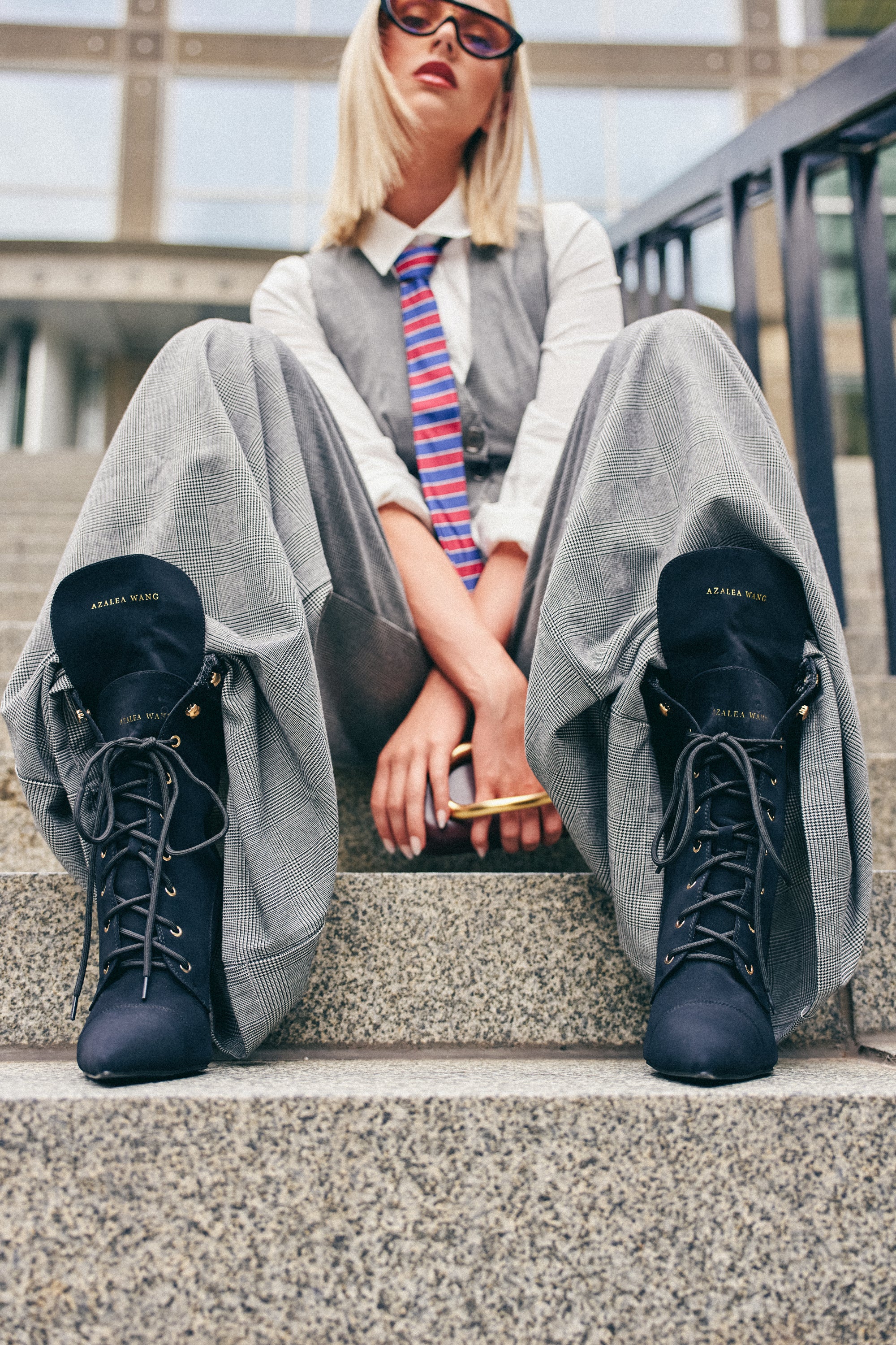 A person in wide gray pants, a white shirt, vest, colorful striped tie, and sunglasses sits confidently on outdoor steps holding a pen. Their look is completed by the AZALEA WANG XIOMARAN BLACK STILETTO BOOTIE—black suede booties featuring subtle snakeskin detailing and a sleek stiletto heel.