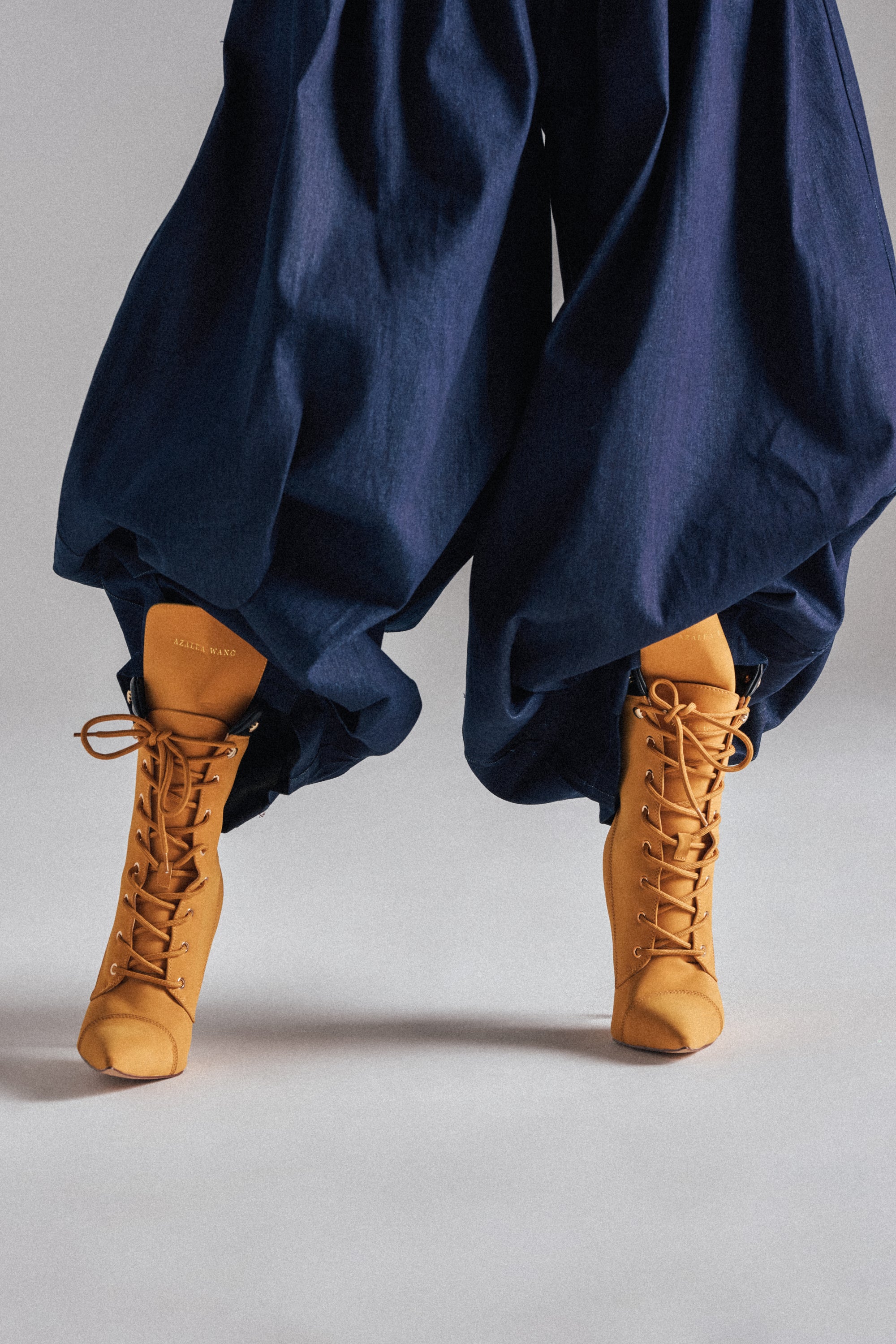 A person stands on tiptoe against a plain light background, showcasing loose navy blue pants paired with the AZALEA WANG XIOMARAN YELLOW BOOTIE, featuring a stiletto heel. The photo focuses on the lower half and their unique combination.