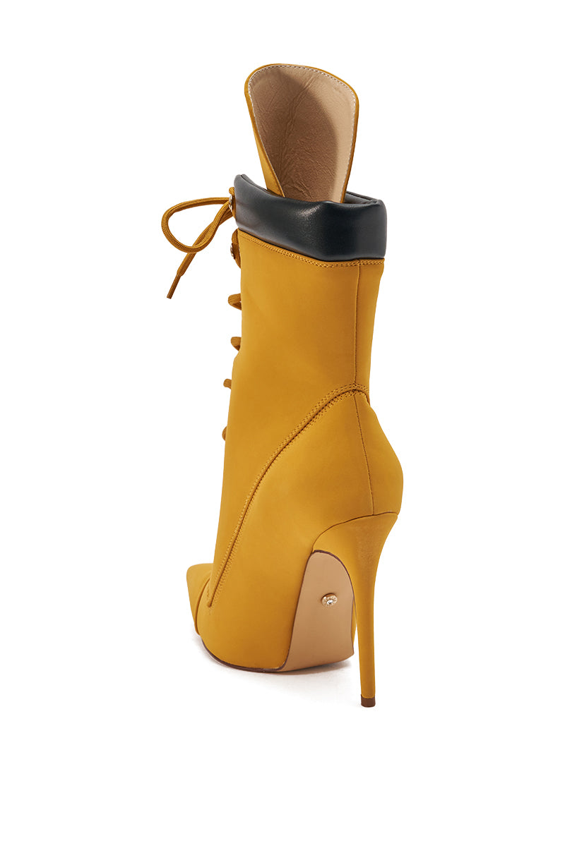 The AZALEA WANG XIOMARAN YELLOW BOOTIE is a mustard yellow faux suede bootie with a pointed toe, stiletto heel, front laces, and black collar, shown from the back on a white background.