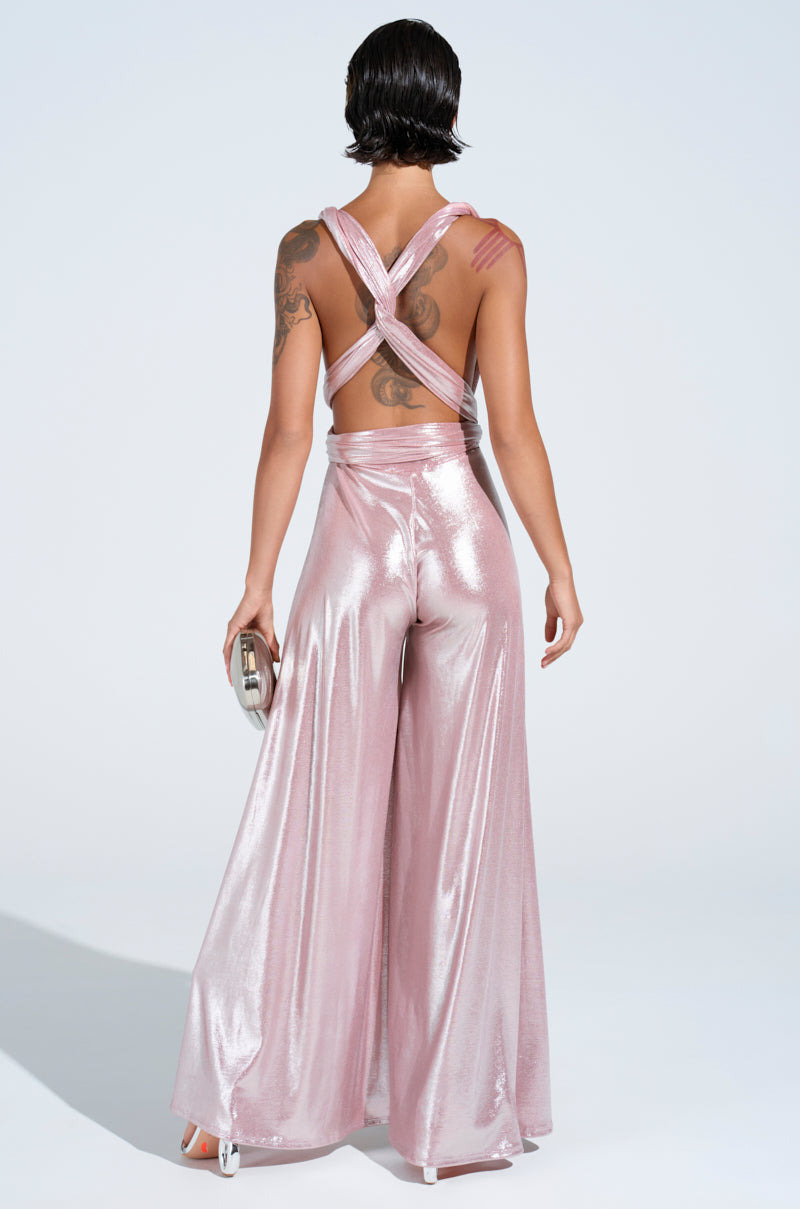 Wearing the SHINE BRIGHT WEAR MULTIPLE WAYS JUMPSUIT in metallic pink with an open back and crisscross straps, a person stands against a light backdrop holding a metallic clutch and wearing white high-heels.