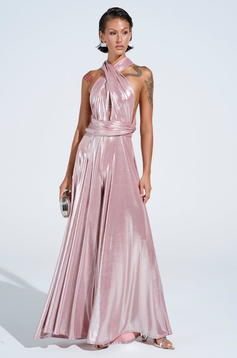 A person stands wearing a shiny rose gold jumpsuit, styled to spotlight its versatile design and self-tie bodice. With a small clutch and strappy silver heels, their short hair and arm tattoos are highlighted against a plain white background.