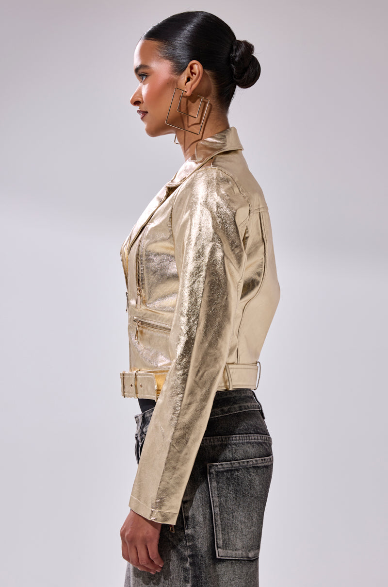 A woman poses in profile wearing the DRIPPING IN GOLD MOTO jacket, black high-waisted jeans, and geometric statement earrings. Her hair is styled in a sleek low bun against a plain light background.