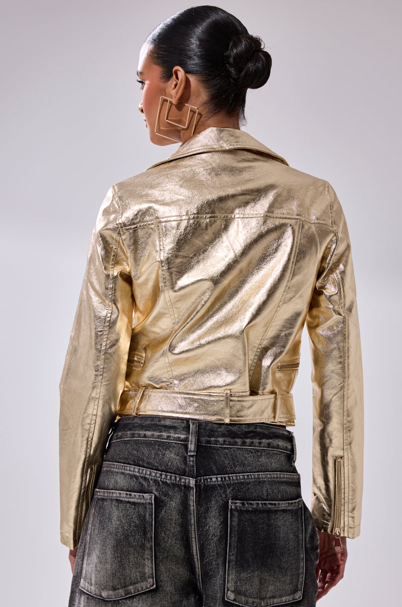 A woman with her hair in a bun, facing away from the camera, models the DRIPPING IN GOLD MOTO jacket and black washed jeans, highlighting her bold geometric earrings against a light gray background.