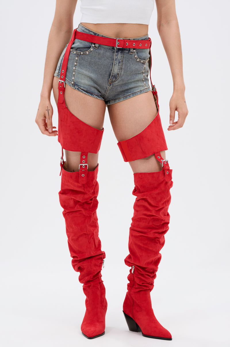 A person stands against a plain light background wearing a white crop top, denim shorts with red belted straps, and the AZALEA WANG XOMARIA RED FAUX SUEDE WESTERN CHAP BOOT—thigh-high with pointed toes and chunky heels. 1