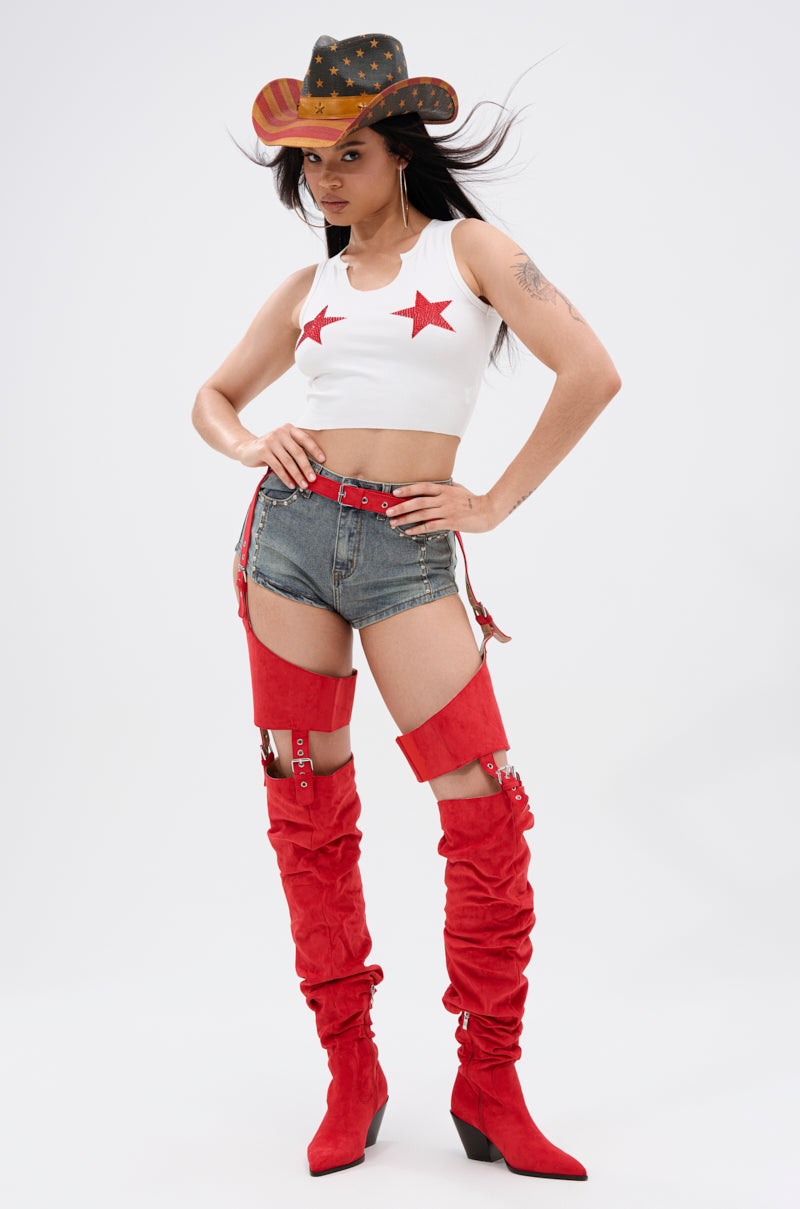 A woman poses confidently in a white crop top with red star pasties, high-waisted denim shorts, the AZALEA WANG XOMARIA RED FAUX SUEDE WESTERN CHAP BOOT, and a black, yellow, and red cowboy hat. 5