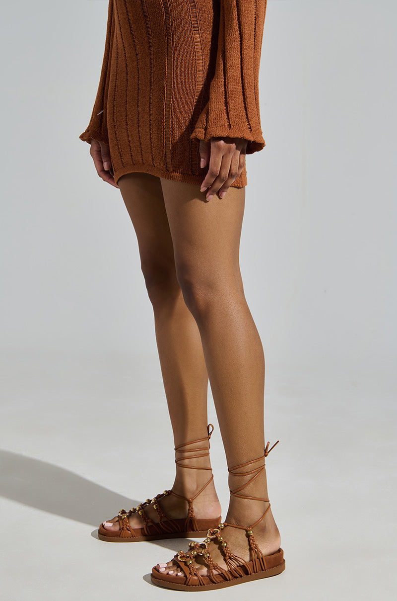 A person in a rust ribbed sweater dress and AZALEA WANG XYLA brown faux suede sandals with metallic gold beads stands against a light gray background. Their relaxed arms highlight the chic outfit and eye-catching footwear.