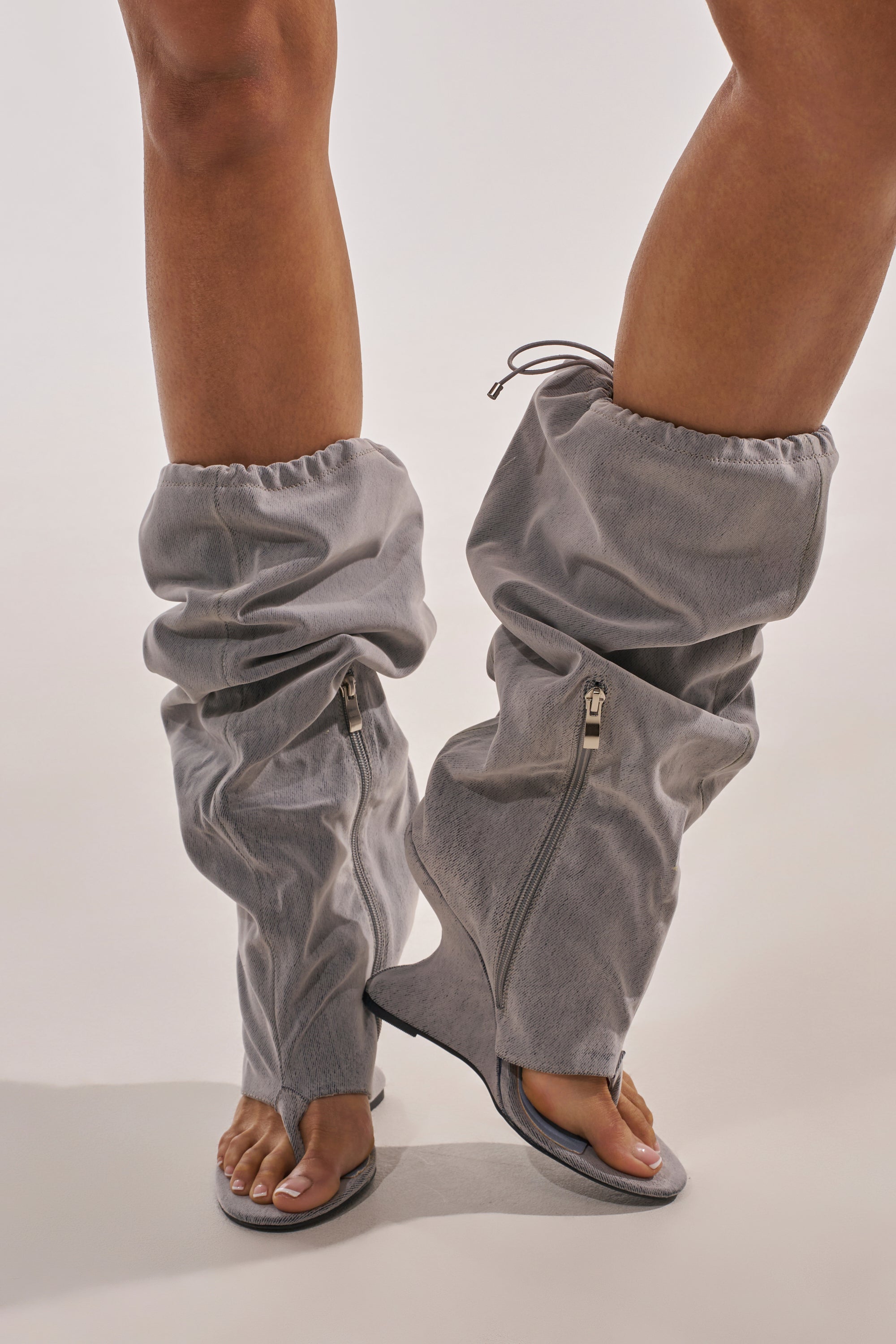 A person wears the AZALEA WANG XYLAH GREY WEDGE GLADIATOR SANDAL—unique gray knee-high sandals with a loose, slouchy fit, open toes and heels, zippers, and drawstrings—standing against a plain light background. 0