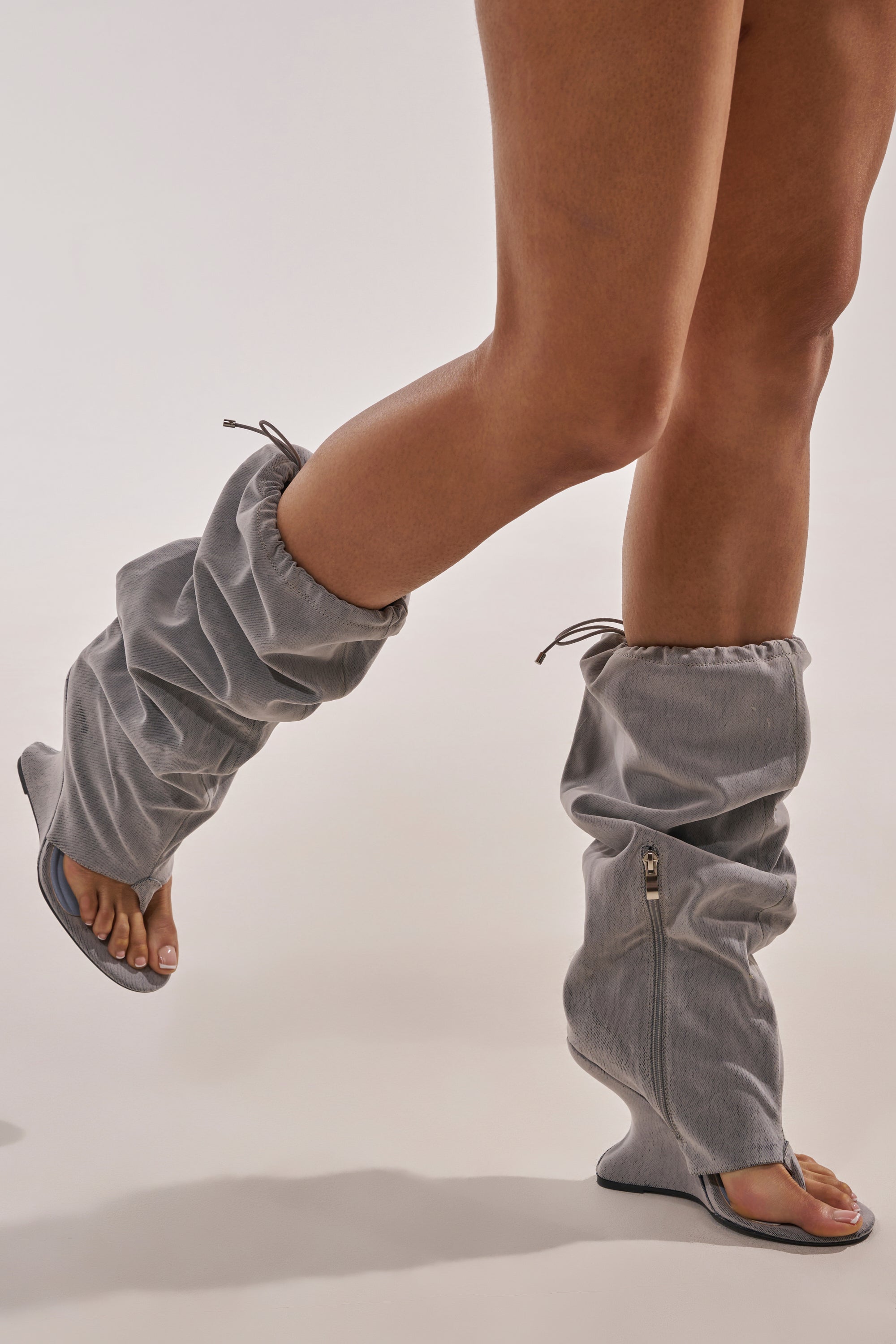 Model wearing the AZALEA WANG XYLAH GREY WEDGE GLADIATOR SANDAL—unique gray knee-high sandals with open toes and heels, ruched fabric, side zippers, and drawstring tops that partially reveal the feet and lower legs. 2