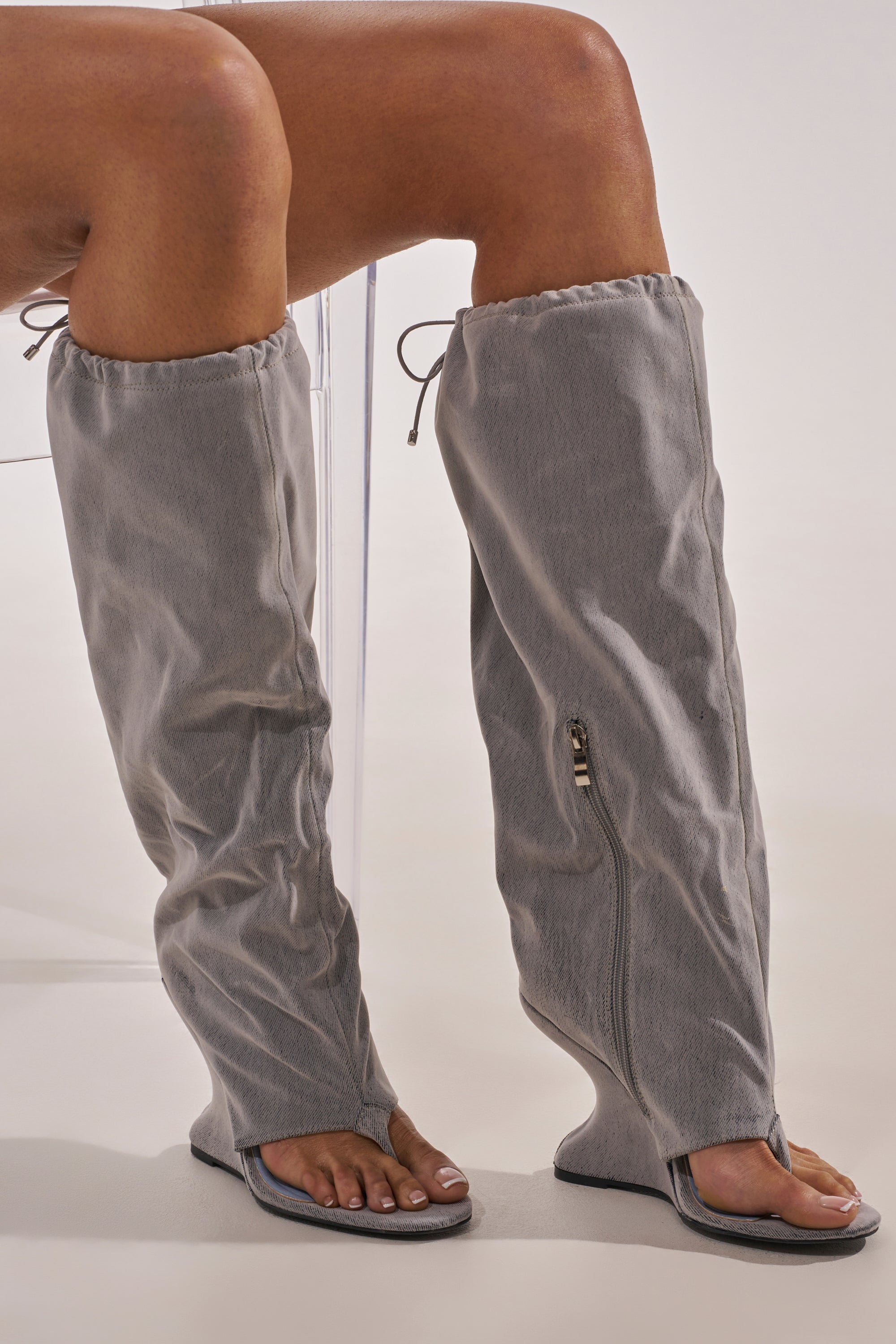 A person is seated, wearing the AZALEA WANG XYLAH GREY WEDGE GLADIATOR SANDAL—unique gray knee-high sandals with open toes, a side zipper, and drawstring tops that blend sandal and boot styles for bold leg coverage. 4