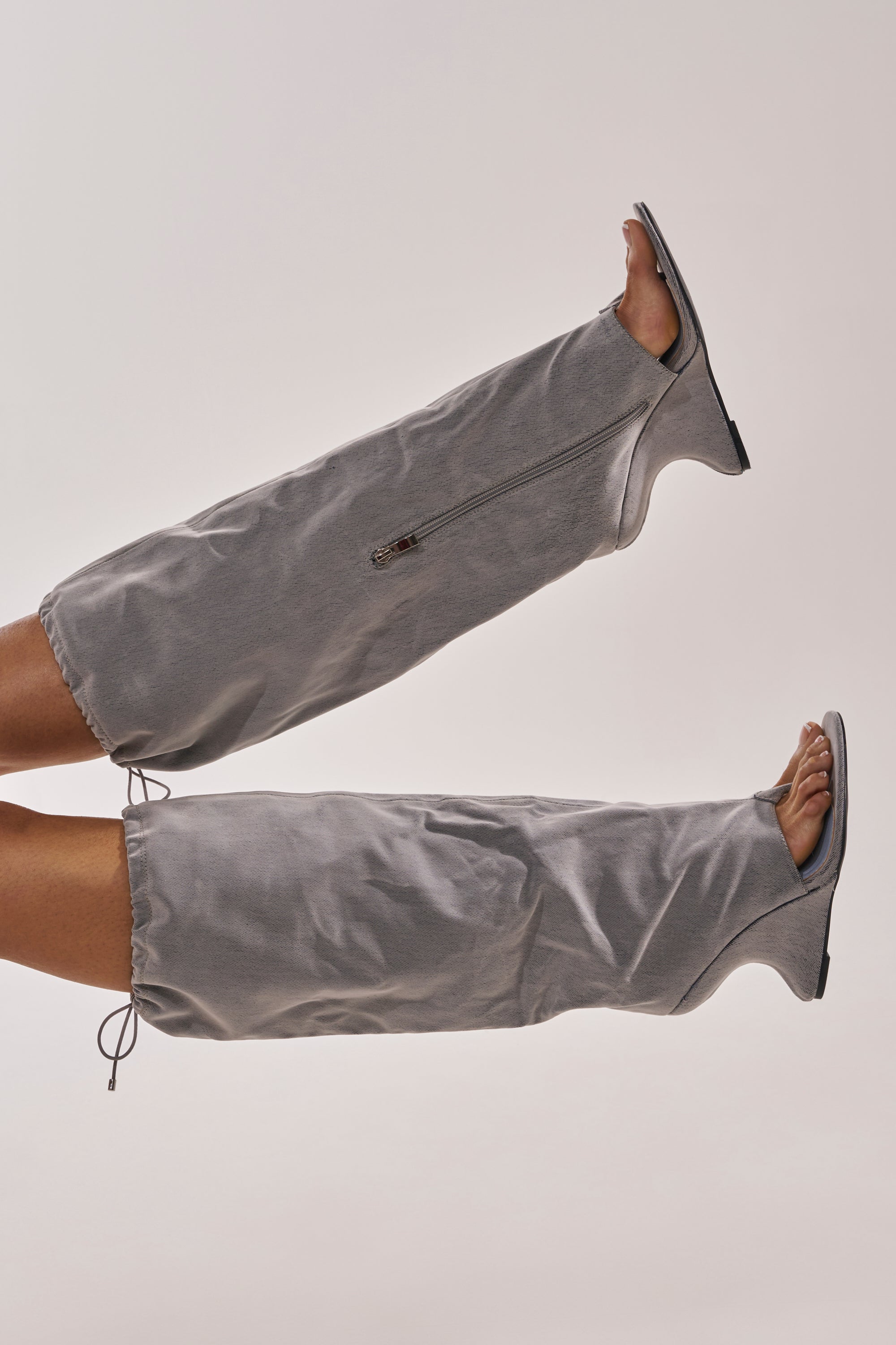 Two legs are raised, wearing AZALEA WANG XYLAH GREY WEDGE GLADIATOR SANDALS with zippers, open toes and heels, plus a loose gray fabric design covering the lower legs, set against a plain light background. 5