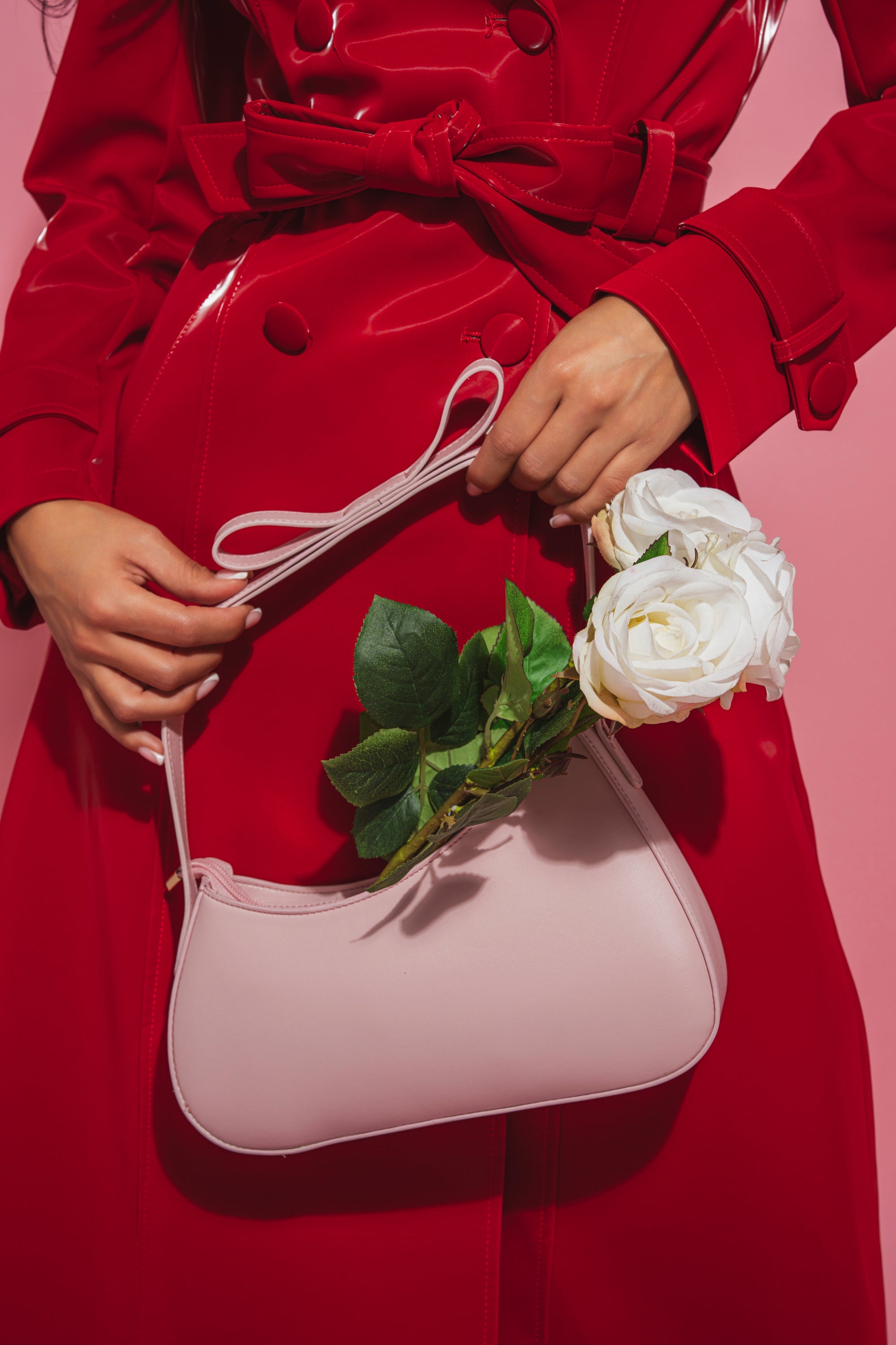 A person in a shiny red trench coat holds the LOVE YOU LIKE A LOVE SONG PURSE, a pale pink faux leather bag filled with two white roses, against a pink background. Only their torso and hands are visible. 1