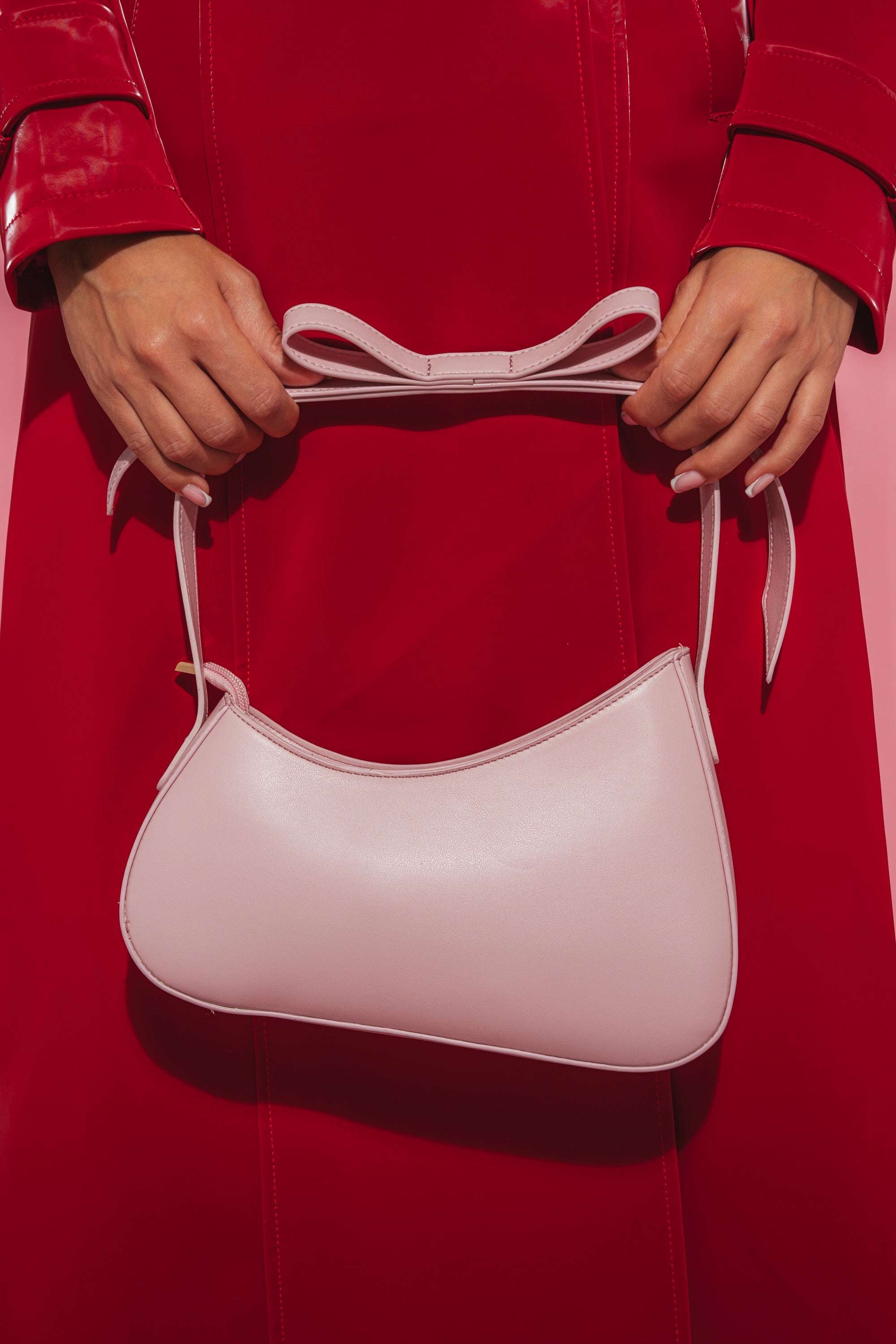 A person in a shiny red coat holds the LOVE YOU LIKE A LOVE SONG PURSE, a pink faux leather bag with a bow-shaped strap, against a matching red background. 2