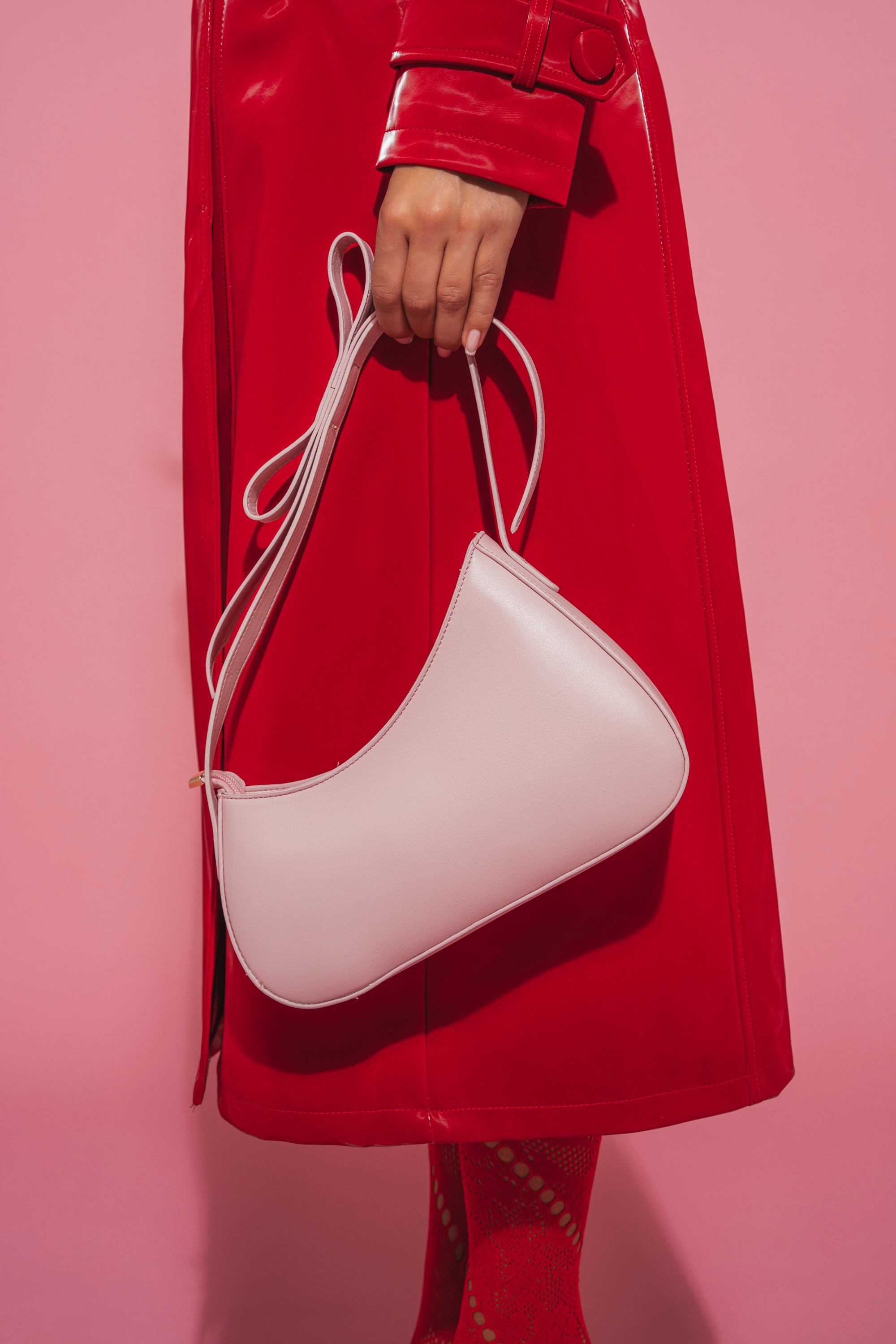 A person in a shiny red coat holds the LOVE YOU LIKE A LOVE SONG PURSE, a pale pink crescent handbag with a long strap, against a pink background; only the hand, bag, and part of the coat are visible. 6