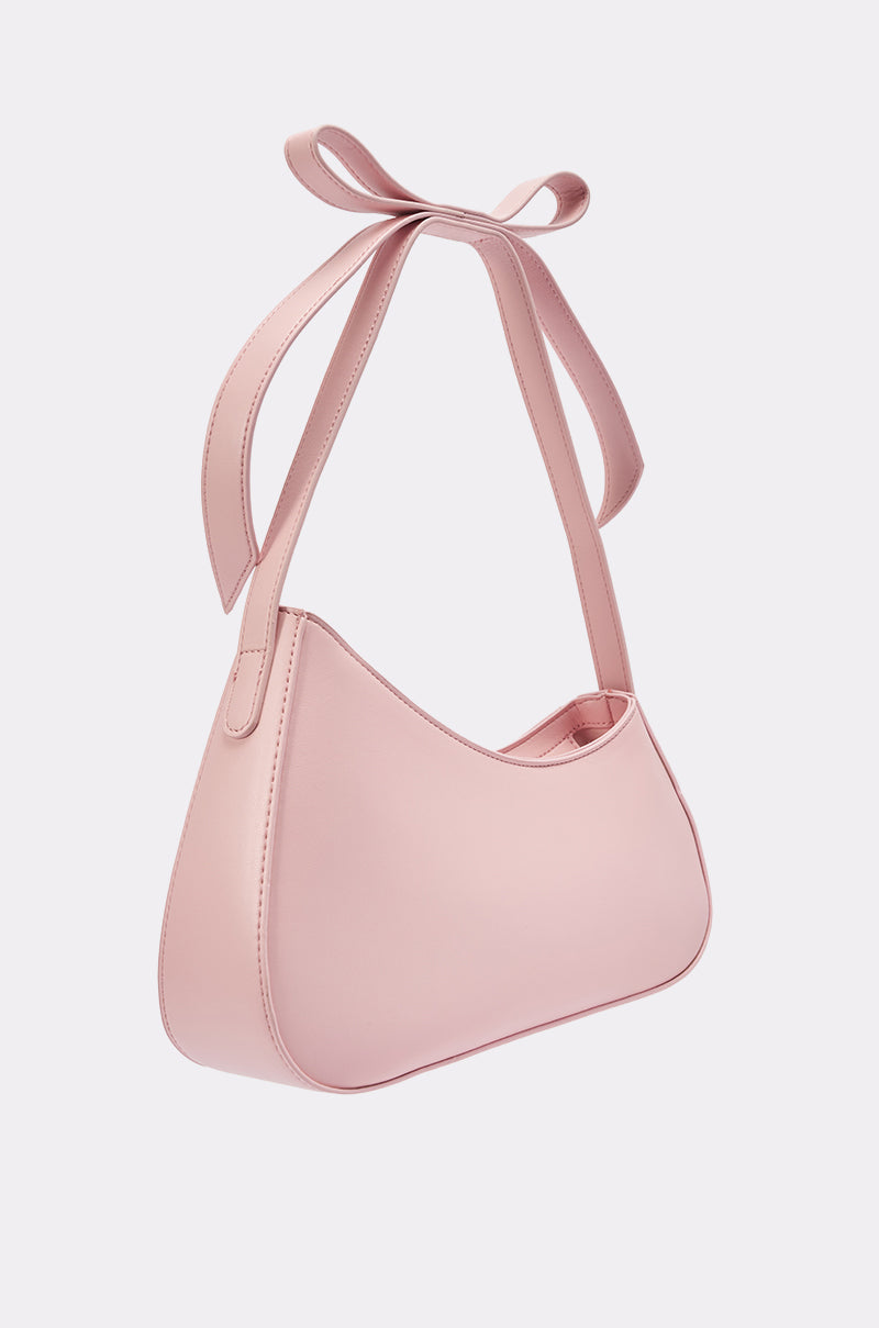 The LOVE YOU LIKE A LOVE SONG PURSE is a light pink faux leather bag with a smooth finish, a chic crescent shape, and a unique top strap tied in a bow, shown against a plain white background. 4