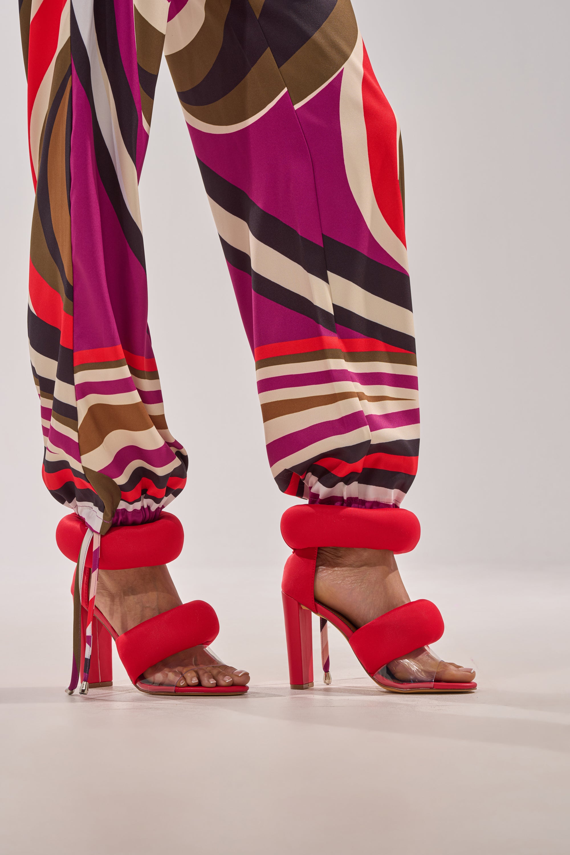 A person wears bold, patterned pants in pink, red, brown, and white tones with the YANIX RED PUFF SLIP ON SANDAL—featuring puffy ankle straps and a cushioned insole—against a plain background. 1