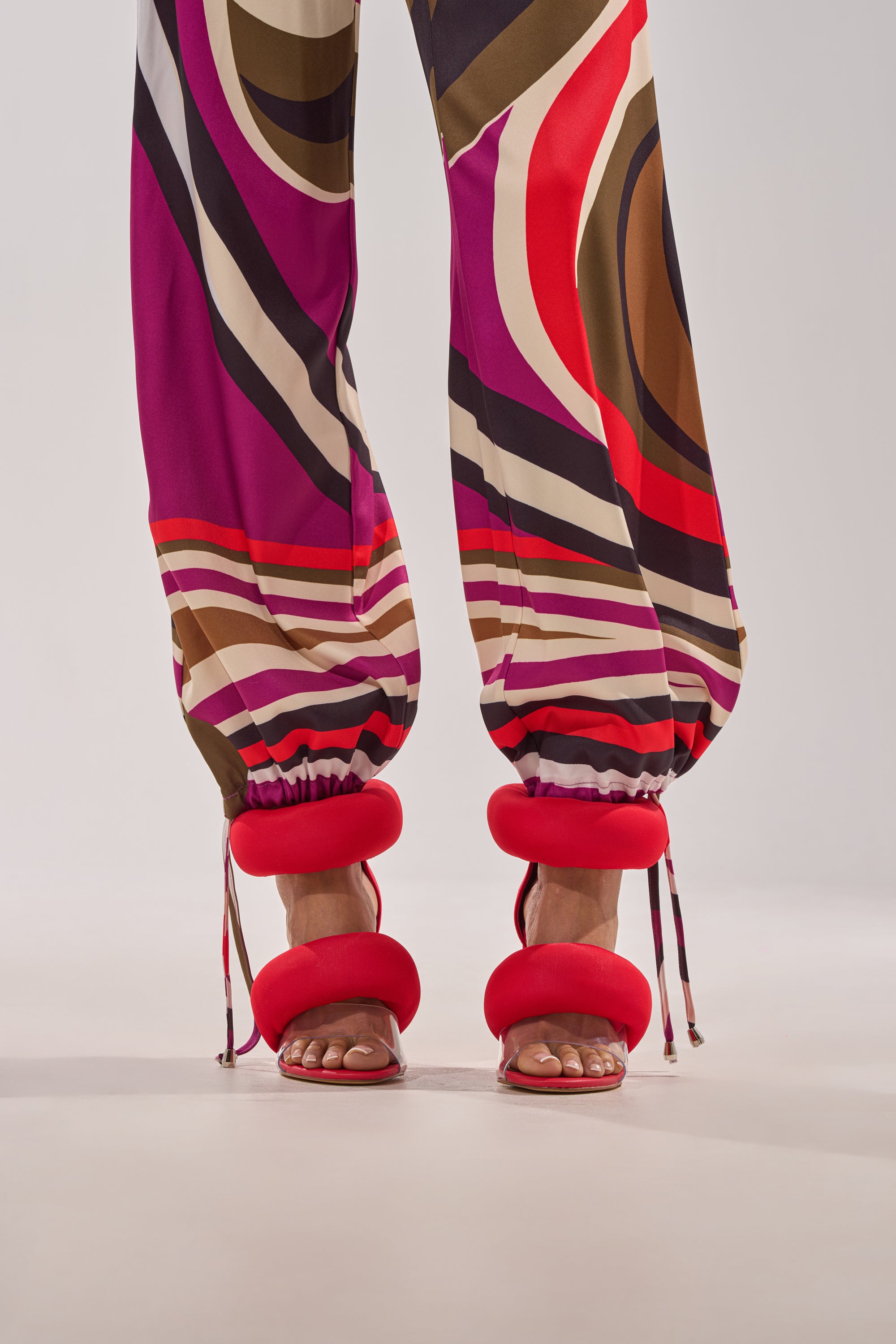 A person wearing colorful, retro-patterned pants and the YANIX RED PUFF SLIP ON SANDAL, featuring bold red chunky heels with large circular padded straps and a cushioned insole, stands on a light background. 2