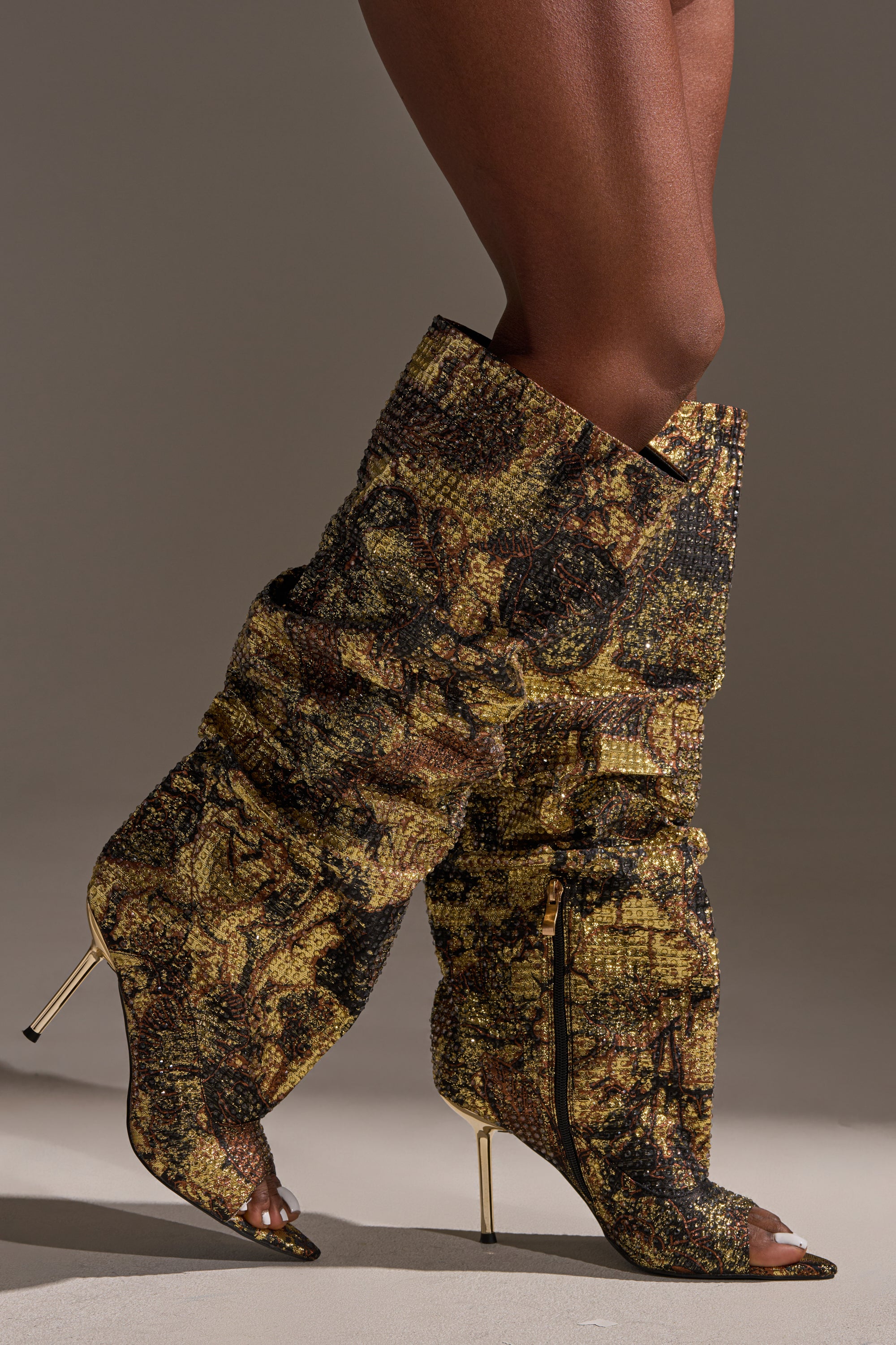 Someone stands on a neutral gray surface wearing the AZALEA WANG YARDLEY MULTI GOLD RHINESTONE COVERED OPEN TOE BOOT—knee-high, pointed-toe boots with a shimmering gold and black pattern that add bold glamour to the look.
