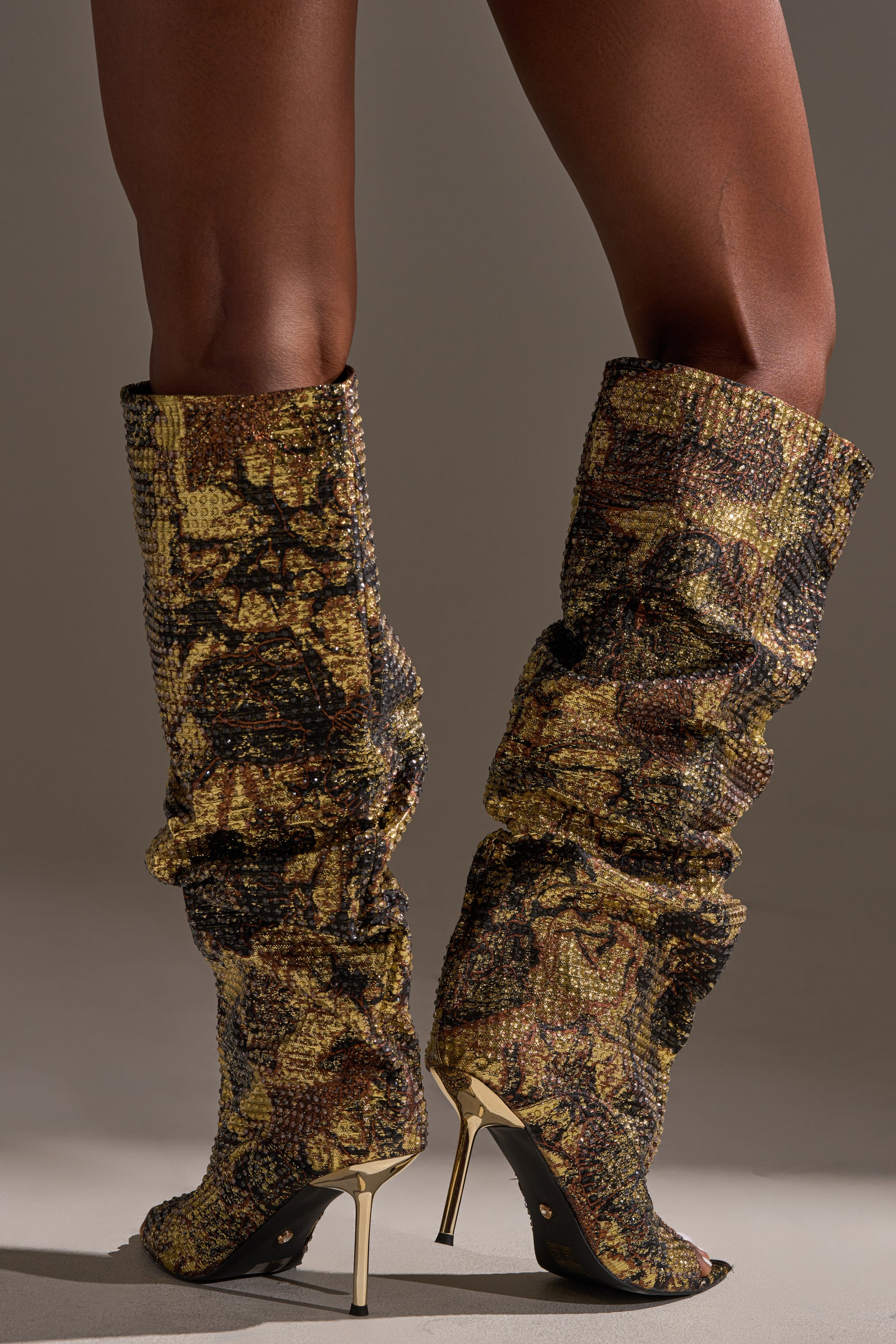 Someone stands on a plain background wearing the AZALEA WANG YARDLEY MULTI GOLD RHINESTONE COVERED OPEN TOE BOOT, featuring tall, slouchy, knee-high stiletto heels in a gold and brown snakeskin pattern.
