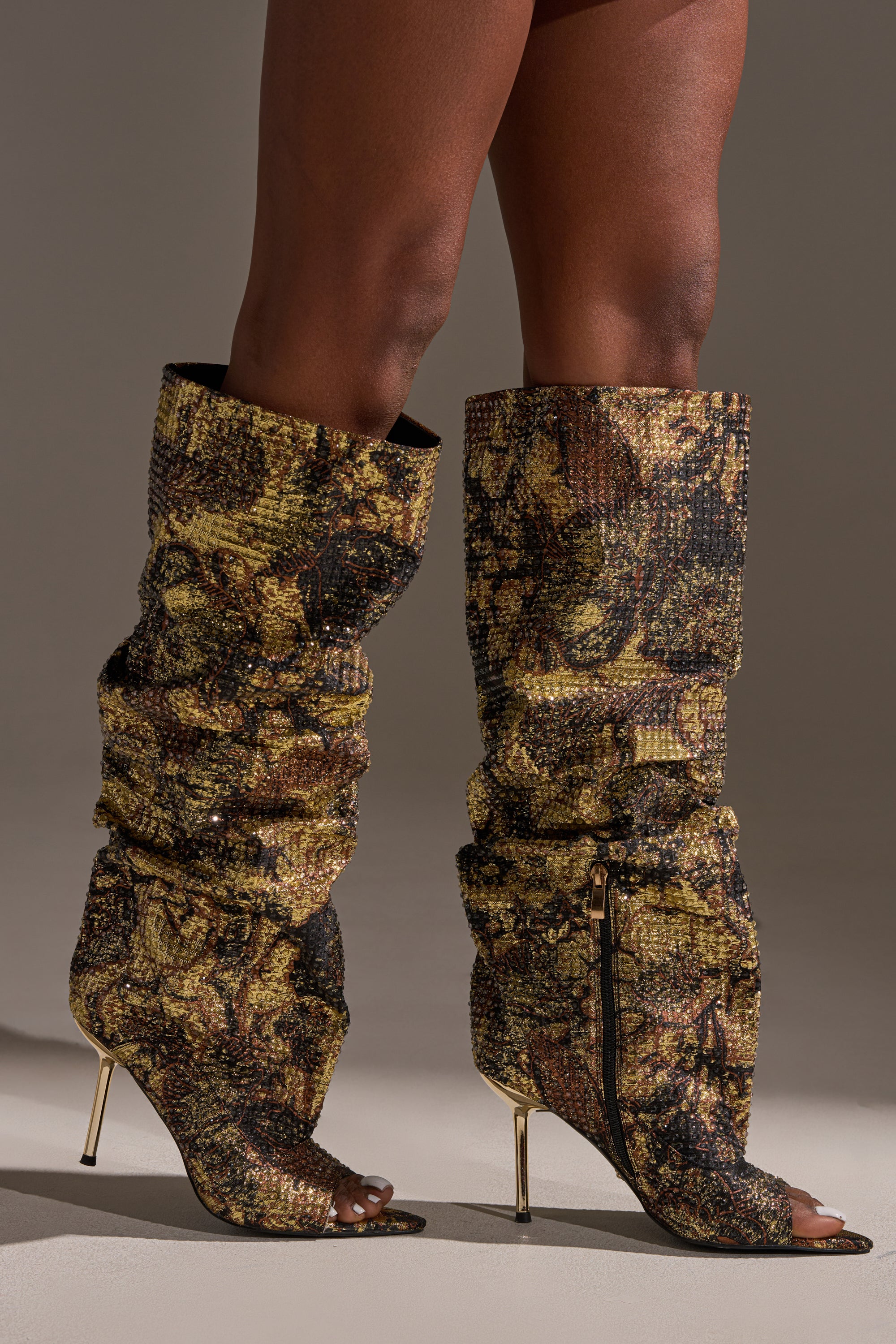 A person wears the AZALEA WANG YARDLEY MULTI GOLD RHINESTONE COVERED OPEN TOE BOOT, featuring tall, slouchy design with gold, brown, and black snakeskin pattern and high thin gold heels, standing on a neutral background.
