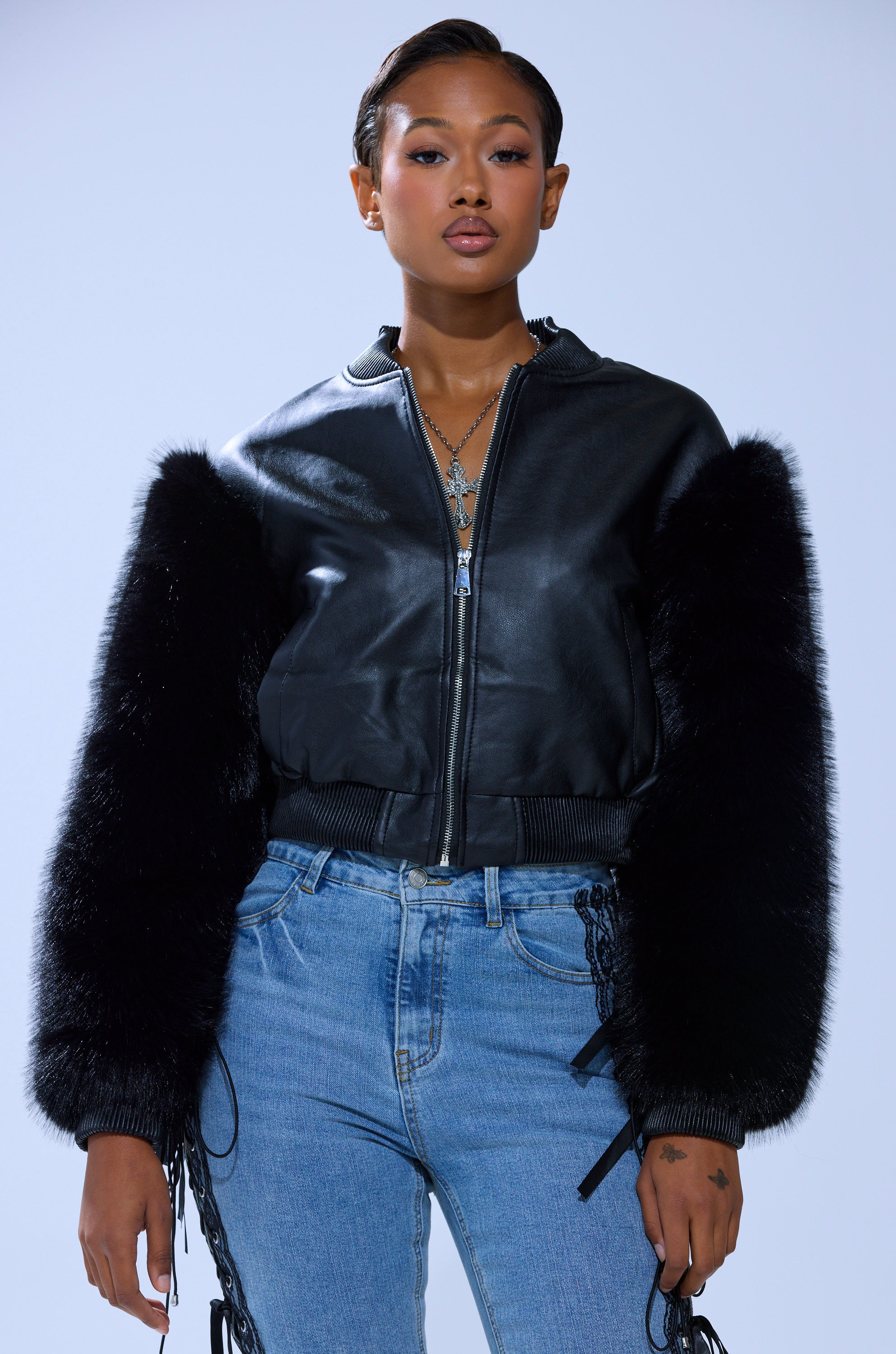 A woman stands confidently in the SO GOOD IT HURTS FUR SLEEVE BOMBER IN BLACK, paired with layered necklaces and blue side-laced jeans against a plain light background.