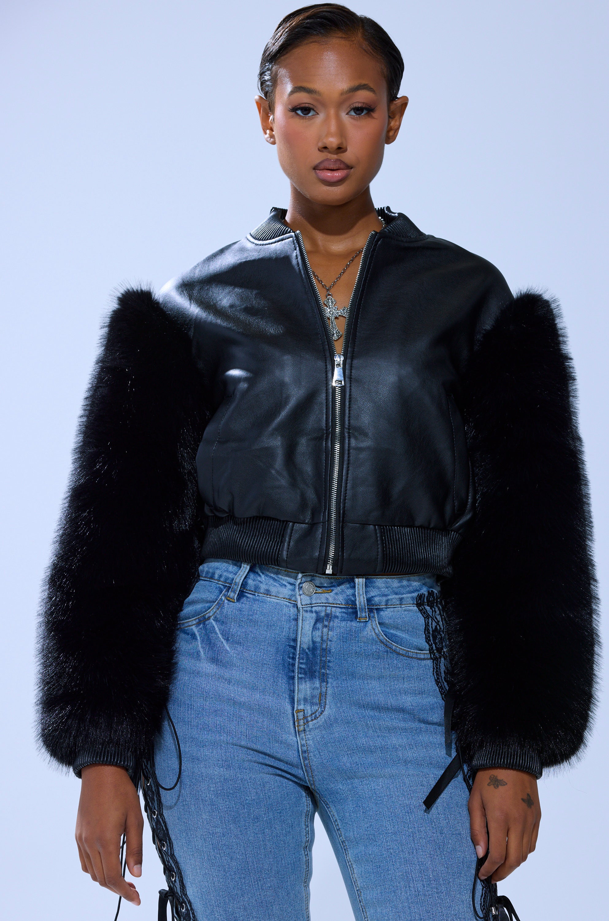 A woman poses confidently against a plain light background wearing the SO GOOD IT HURTS FUR SLEEVE BOMBER IN BLACK, paired with light blue jeans and layered necklaces.