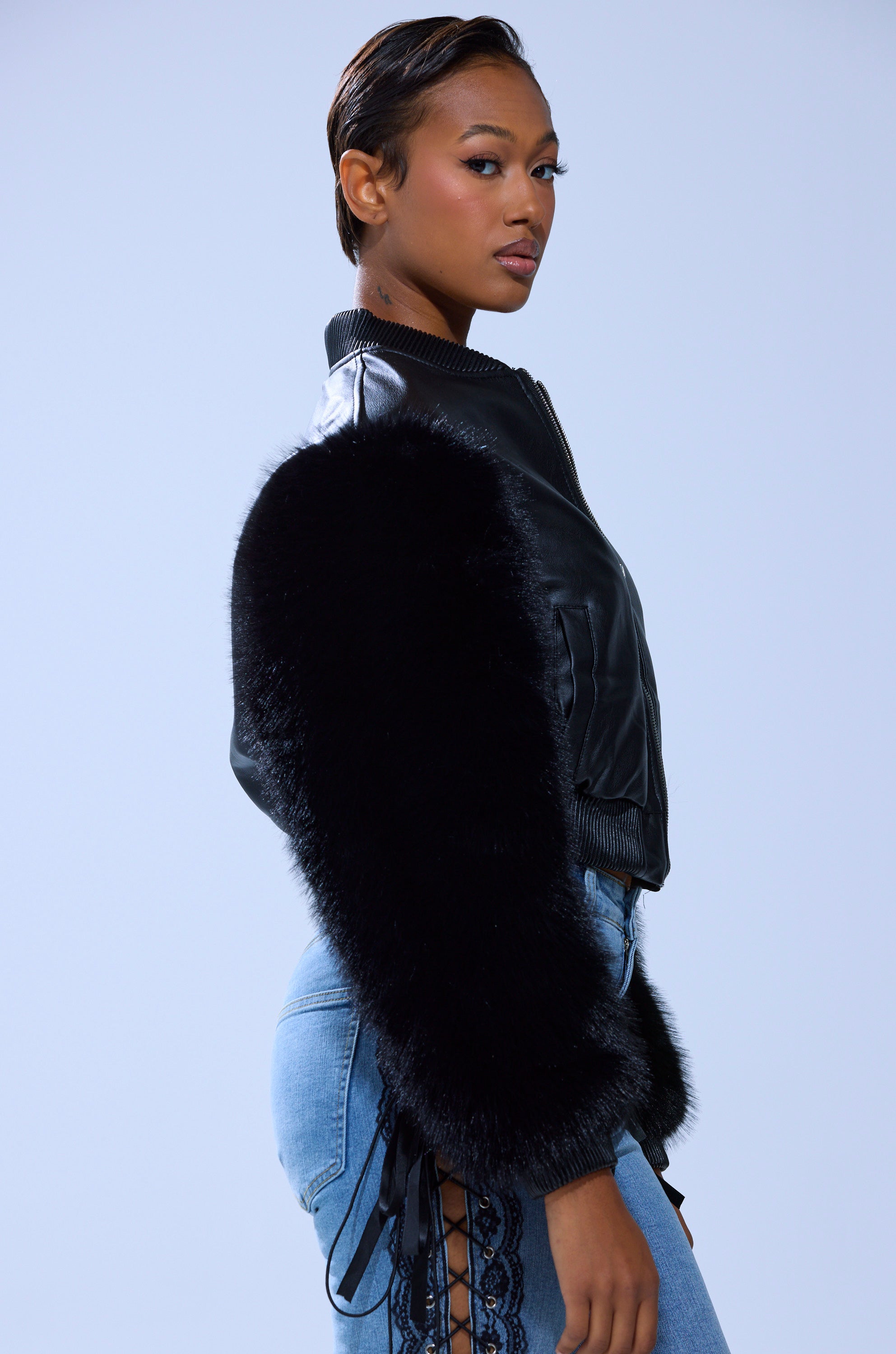 A woman poses sideways in the SO GOOD IT HURTS FUR SLEEVE BOMBER IN BLACK and light blue lace-up jeans, set against a plain light background.