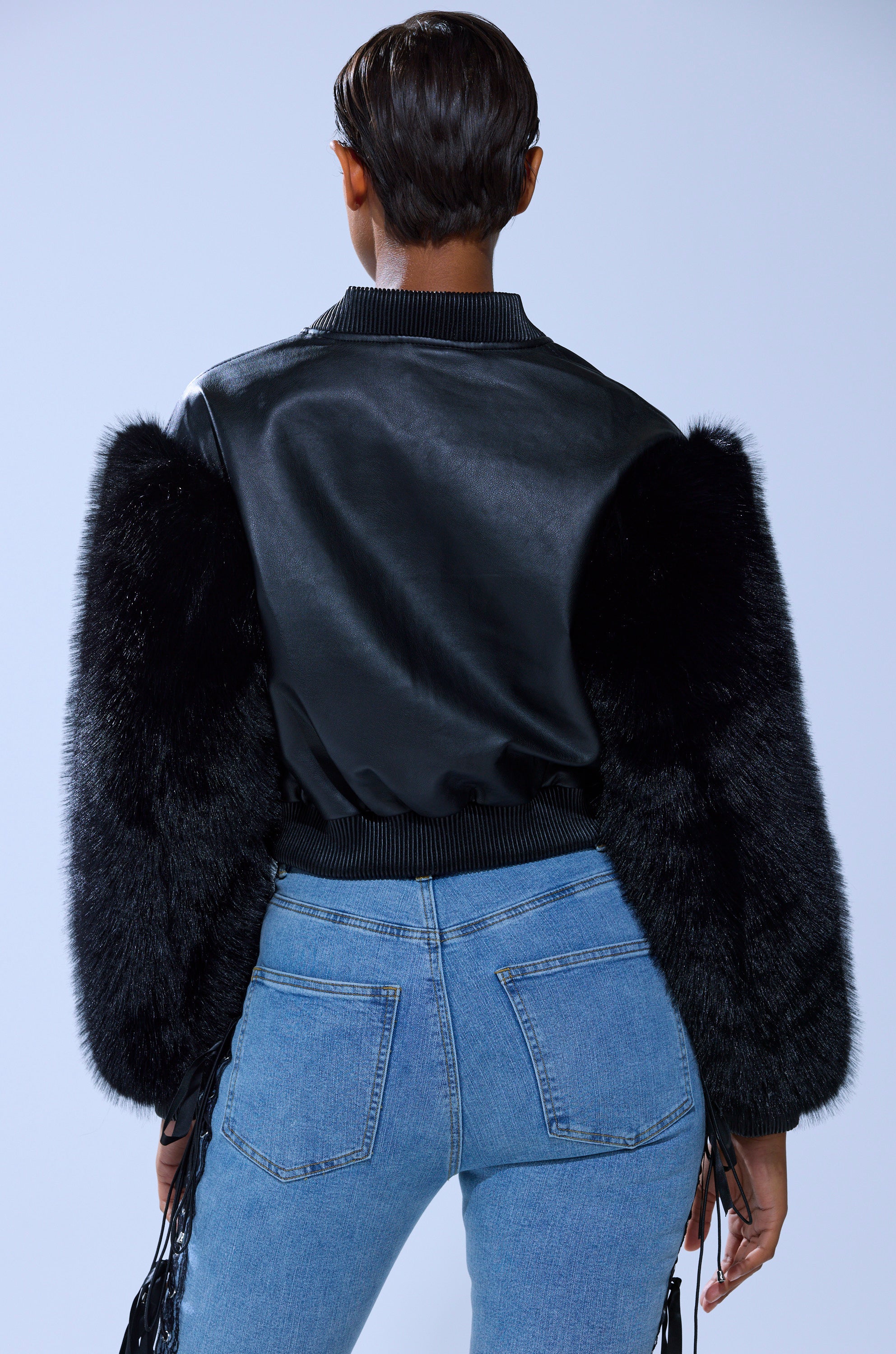 A person with short hair, viewed from behind, wears the SO GOOD IT HURTS FUR SLEEVE BOMBER IN BLACK and light blue jeans, standing against a plain light background.