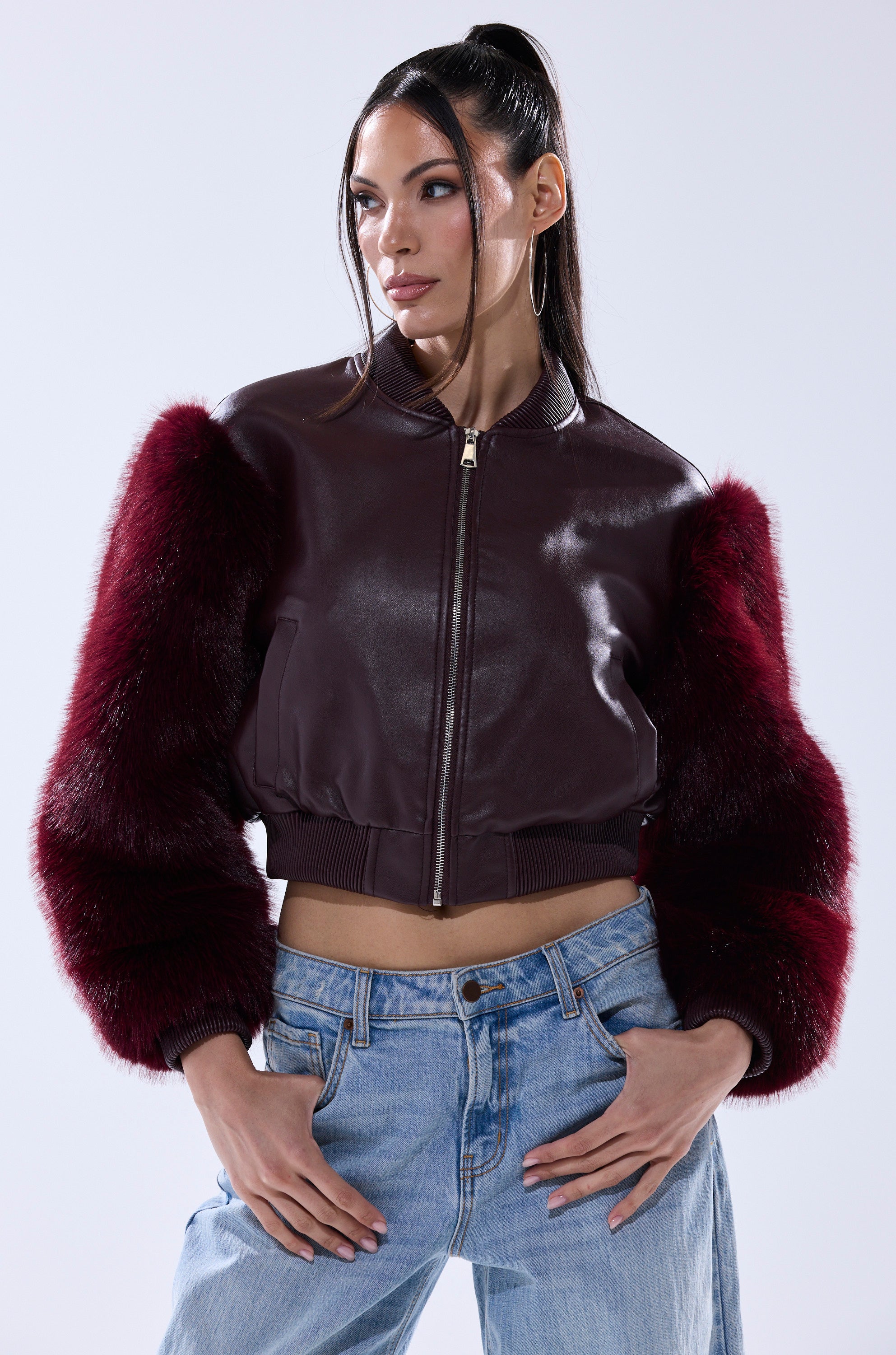 A woman wears the SO GOOD IT HURTS FUR SLEEVE BOMBER IN BURGUNDY, a cropped faux leather jacket with fluffy fur sleeves, paired with light blue jeans. She stands with one hand on her hip, looking to the side.