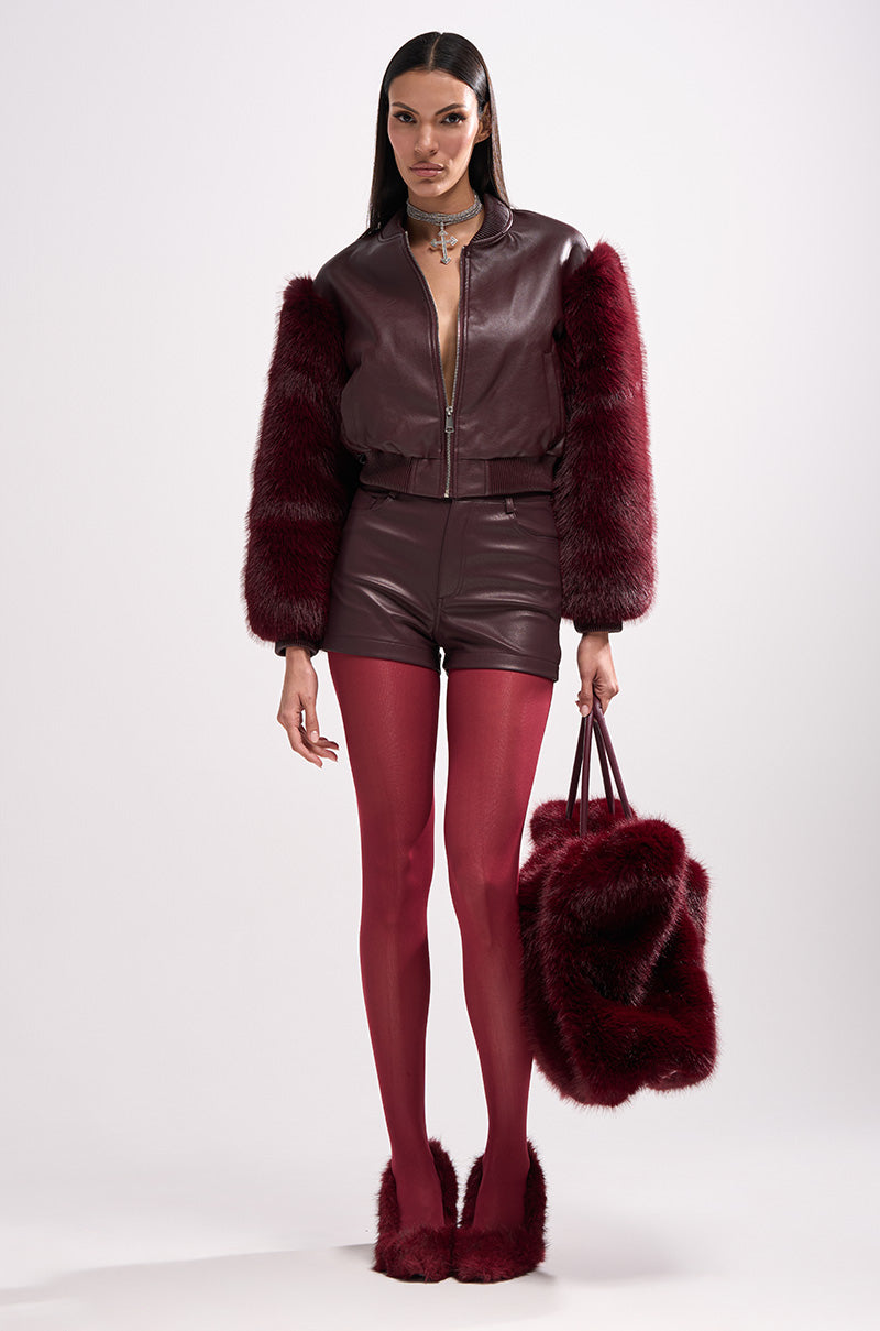 A woman stands against a white background wearing the SO GOOD IT HURTS FUR SLEEVE BOMBER IN BURGUNDY, paired with matching leather shorts, red tights, red fur shoes, and holding a large burgundy faux fur bag.