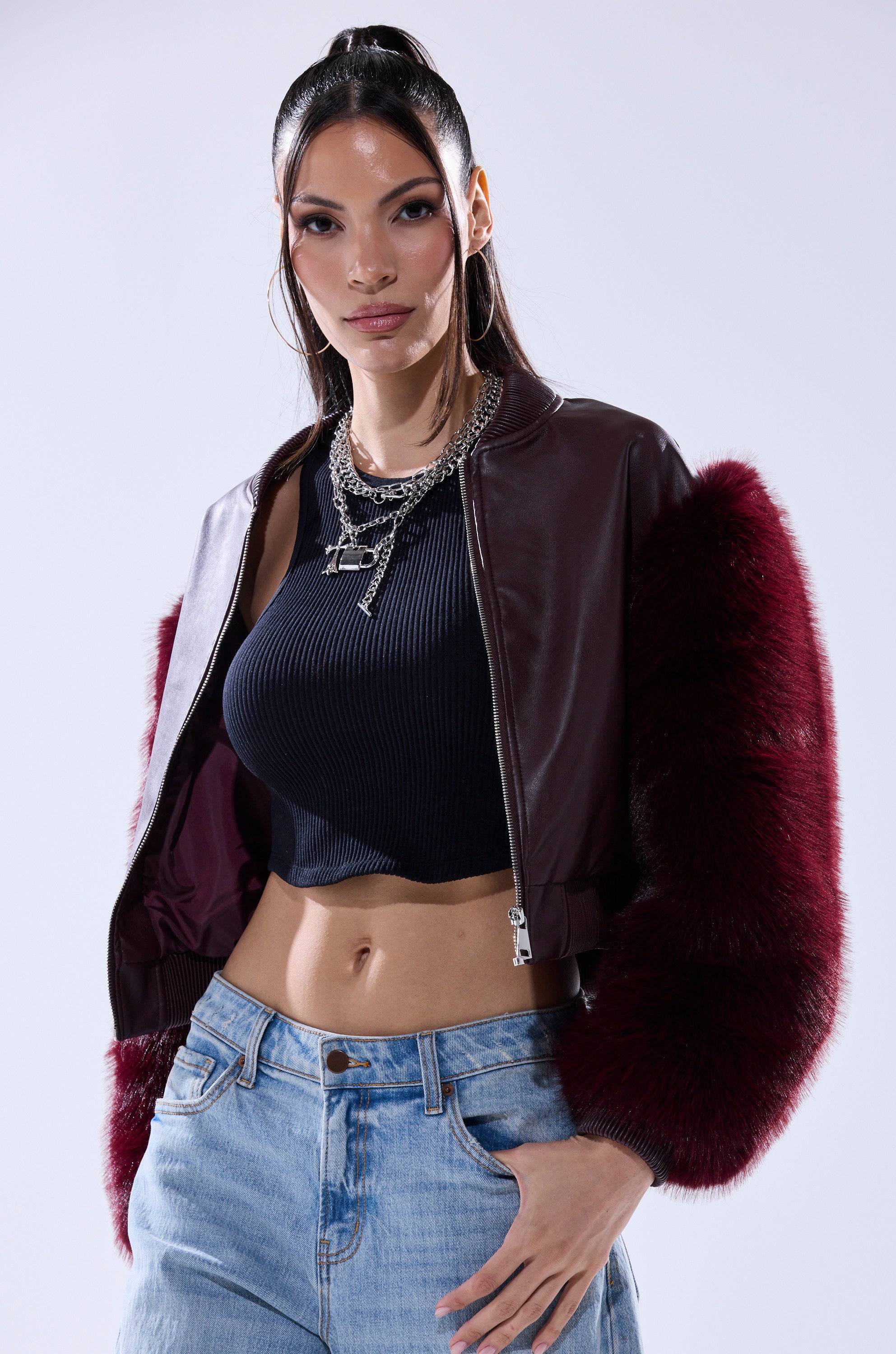 A woman poses confidently in the SO GOOD IT HURTS FUR SLEEVE BOMBER IN BURGUNDY, styled with a black ribbed crop top, layered silver necklaces, and light blue jeans against a simple light background.