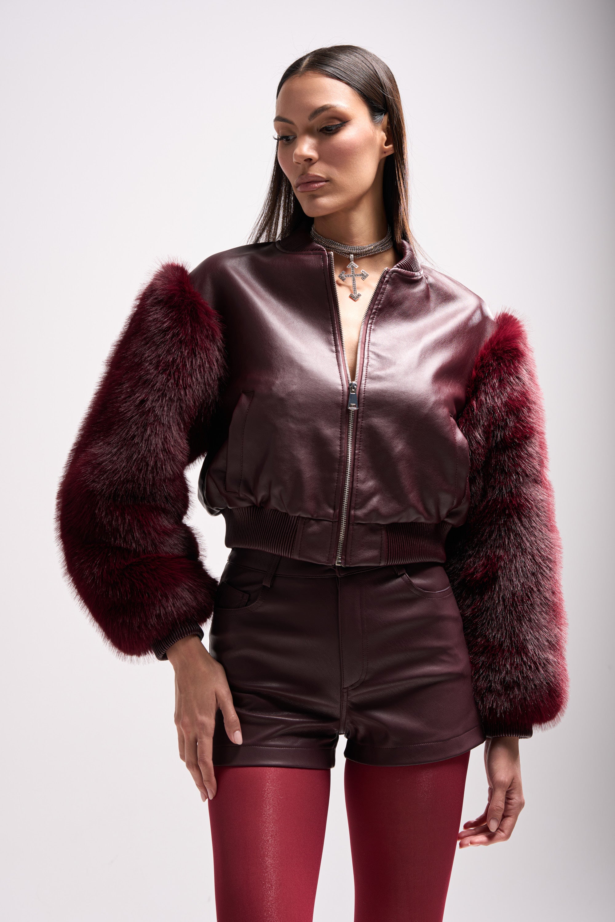 A woman models the SO GOOD IT HURTS FUR SLEEVE BOMBER IN BURGUNDY with faux fur sleeves, paired with matching leather shorts and red tights against a plain light background.