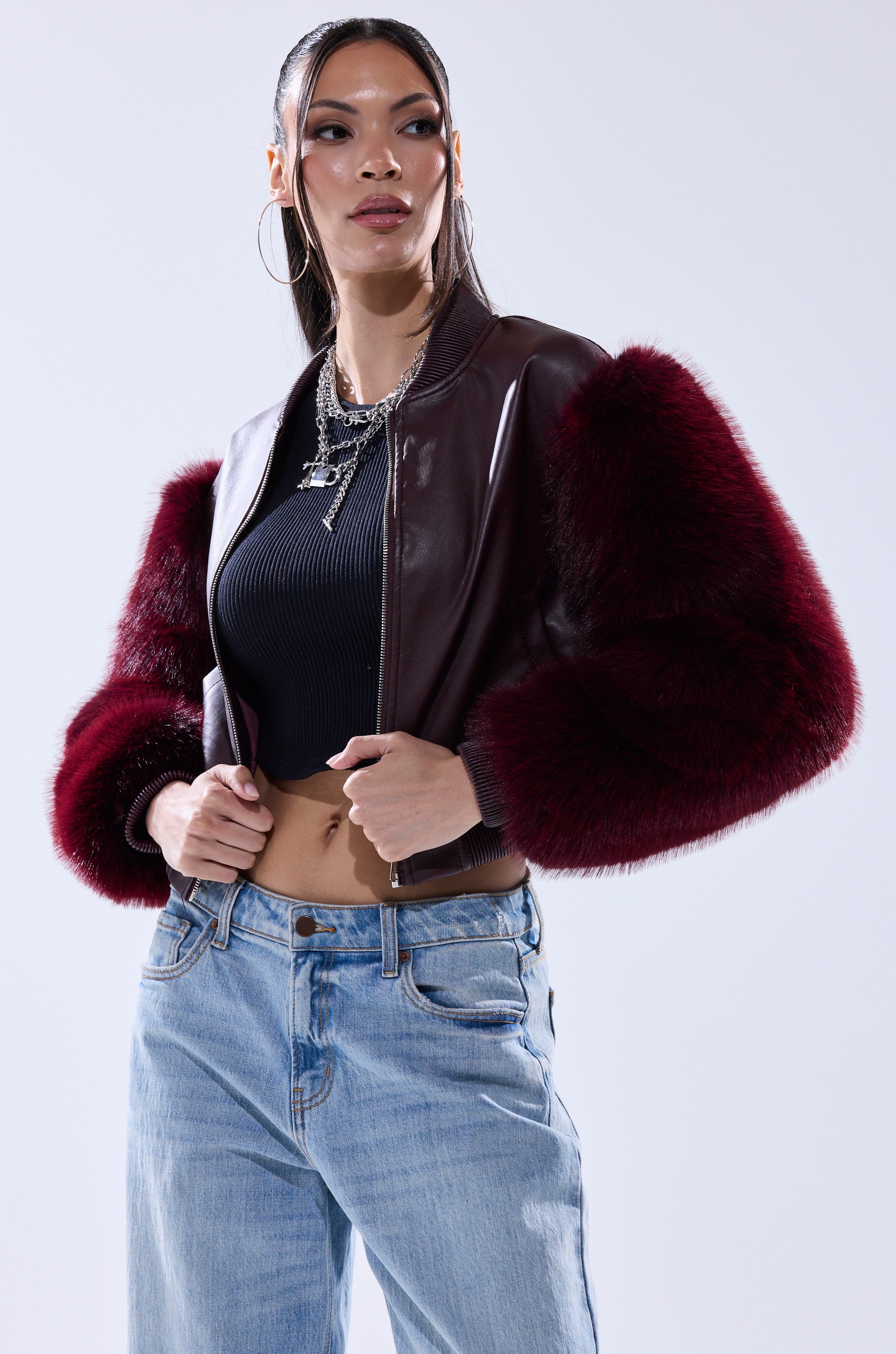 A woman poses confidently in hoop earrings, layered necklaces, a black cropped top, light blue jeans, and the SO GOOD IT HURTS FUR SLEEVE BOMBER IN BURGUNDY against a light background.